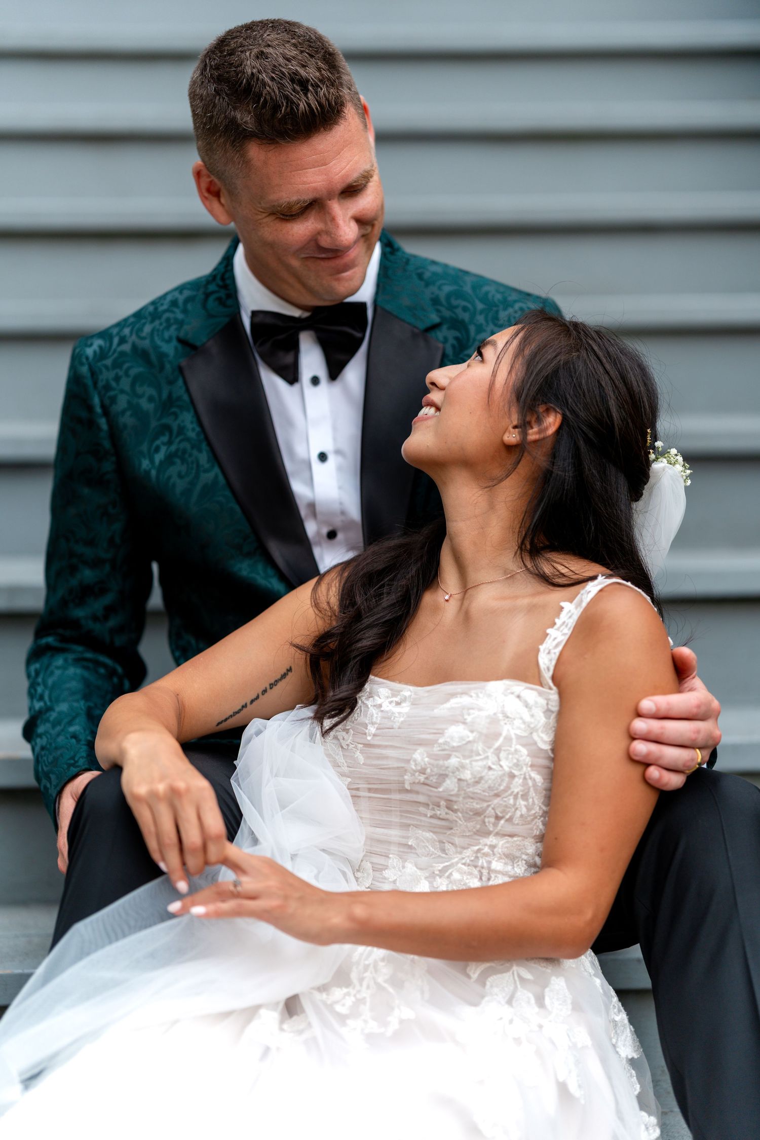Bride in white lace gown sits on steps gazing up at groom in green floral tuxedo jacket with black bow tie.
