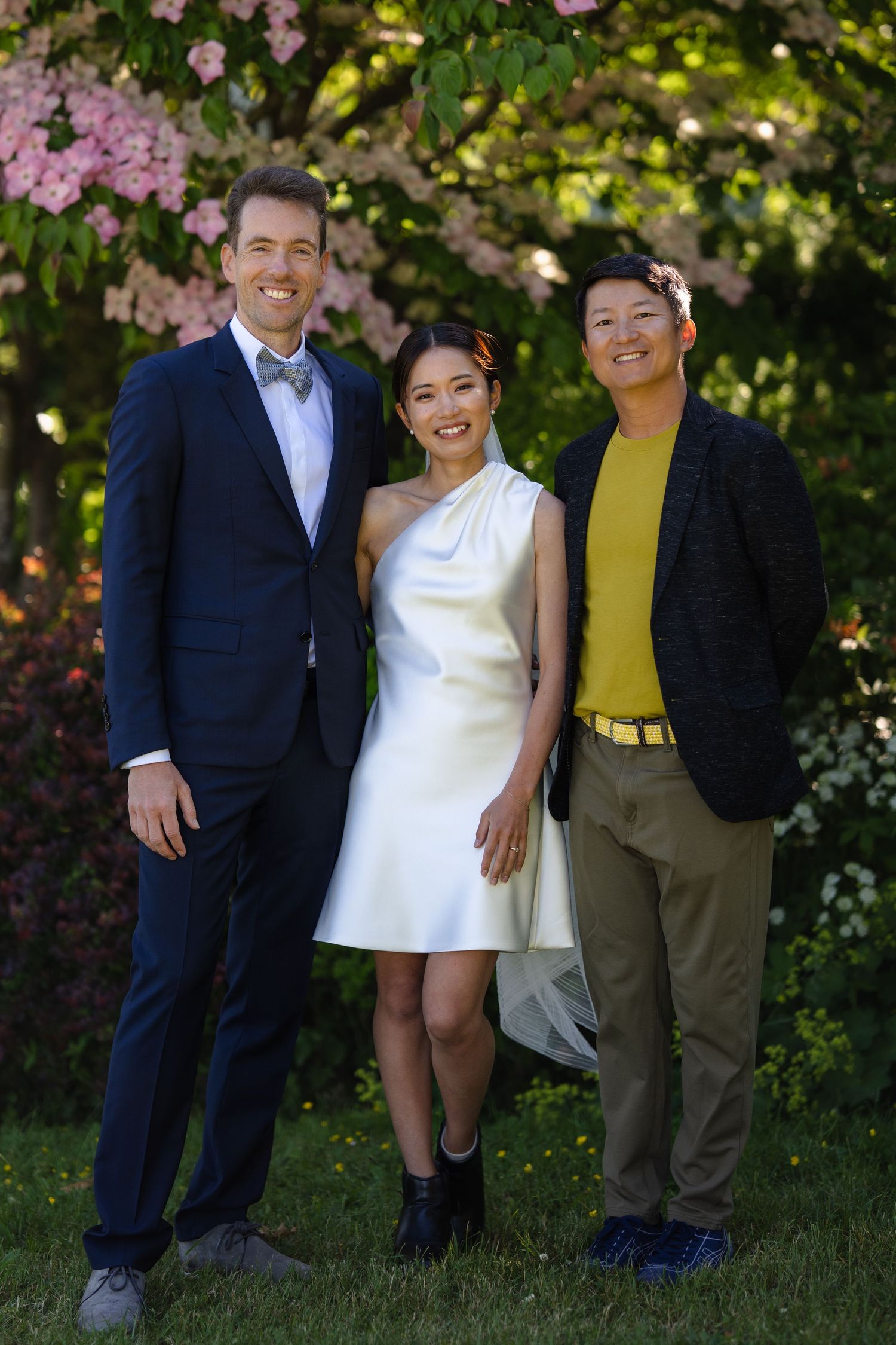 Three smiling people pose outdoors in formal attire near blooming pink flowers on a sunny day.