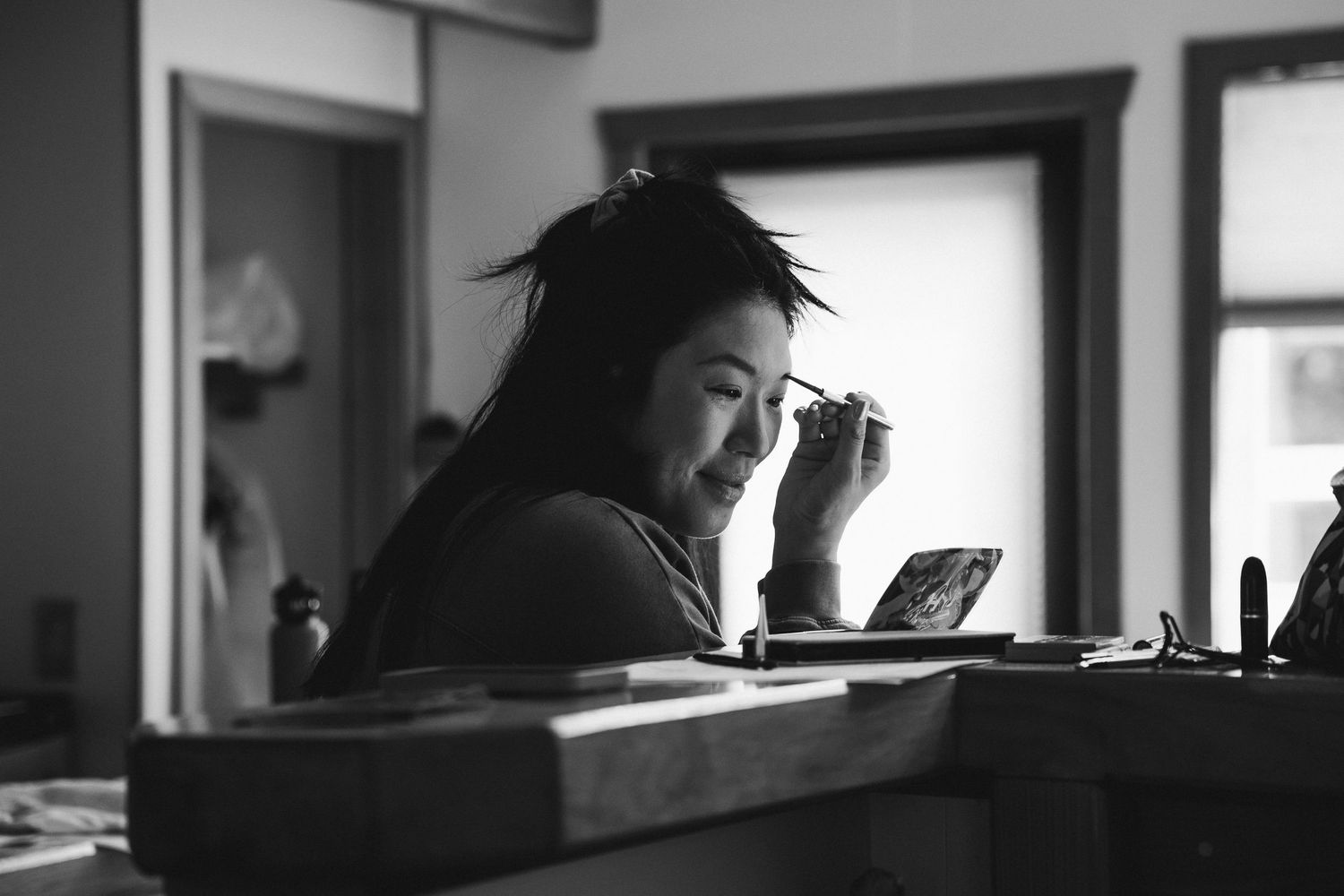 Black and white photo of a young Asian woman applying makeup at a vanity mirror indoors.