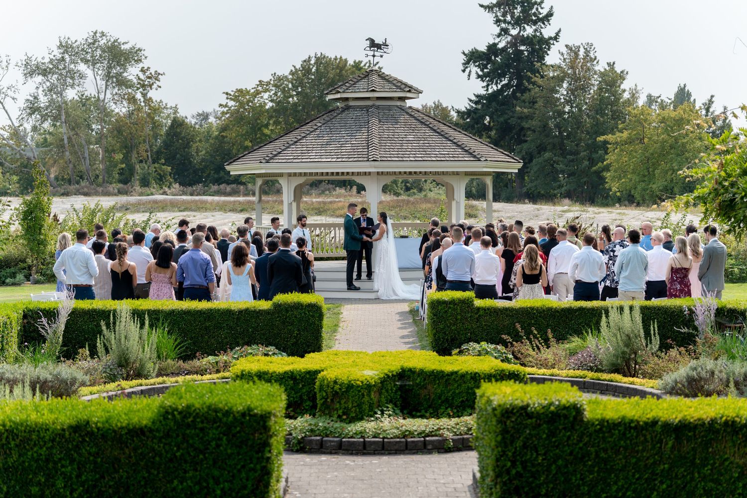 Outdoor wedding ceremony at a garden gazebo surrounded by manicured hedges and guests.