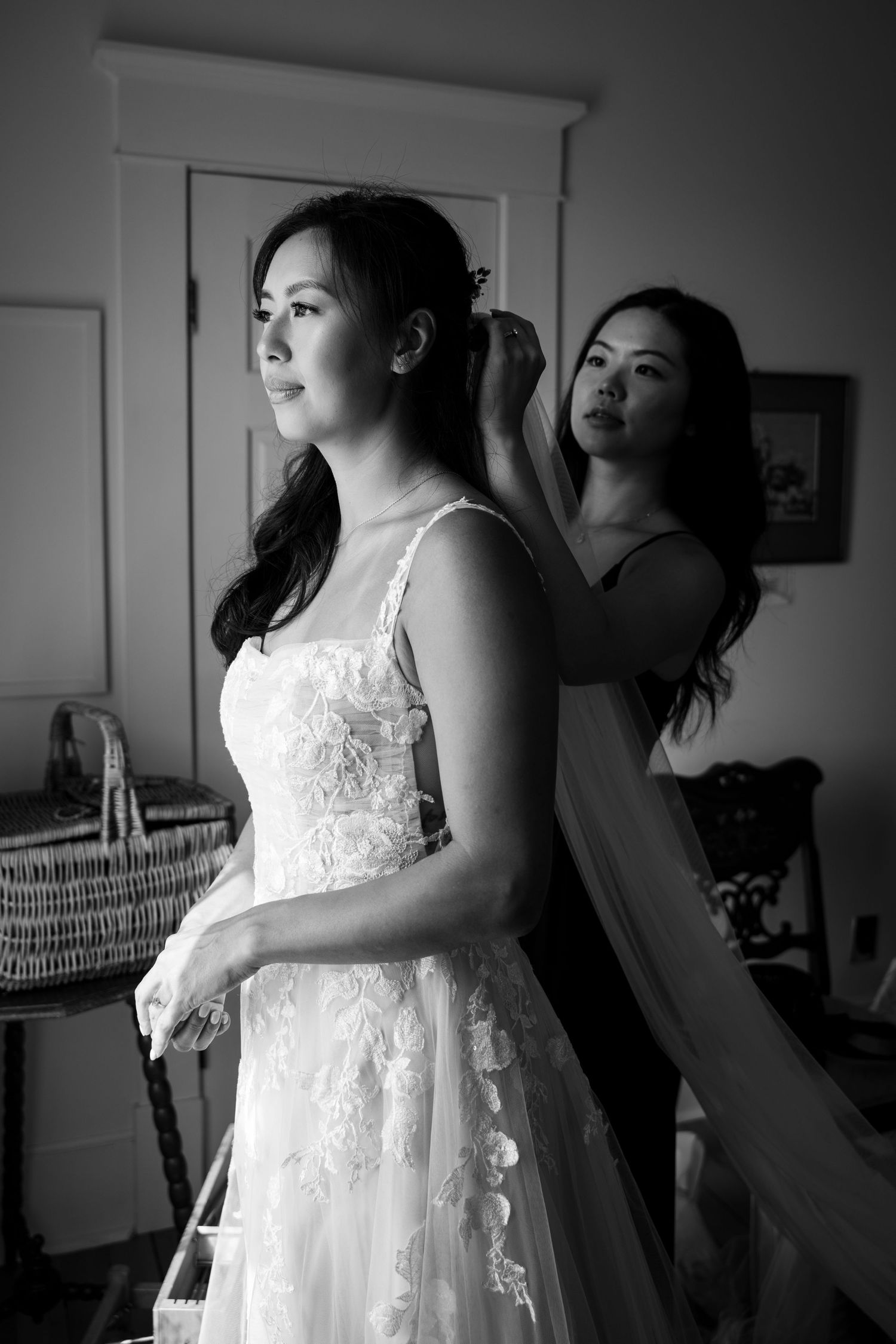 Black and white photo of bride in lace wedding gown having her hair styled by a bridesmaid.