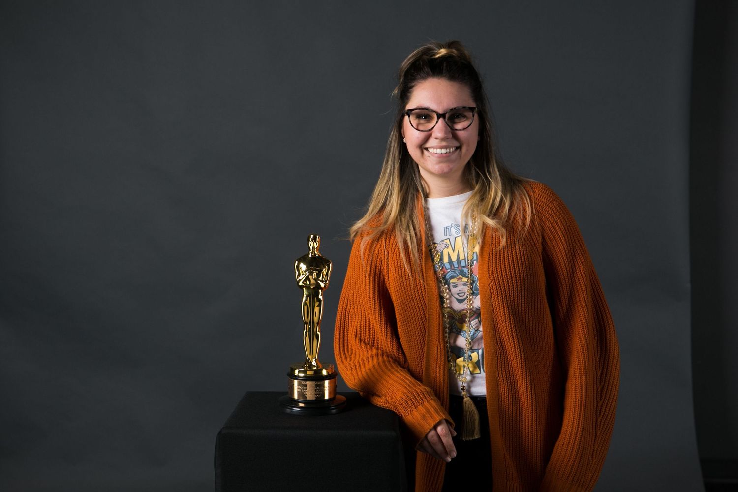 Woman in orange cardigan smiling next to an Oscar award statue on a dark studio background.