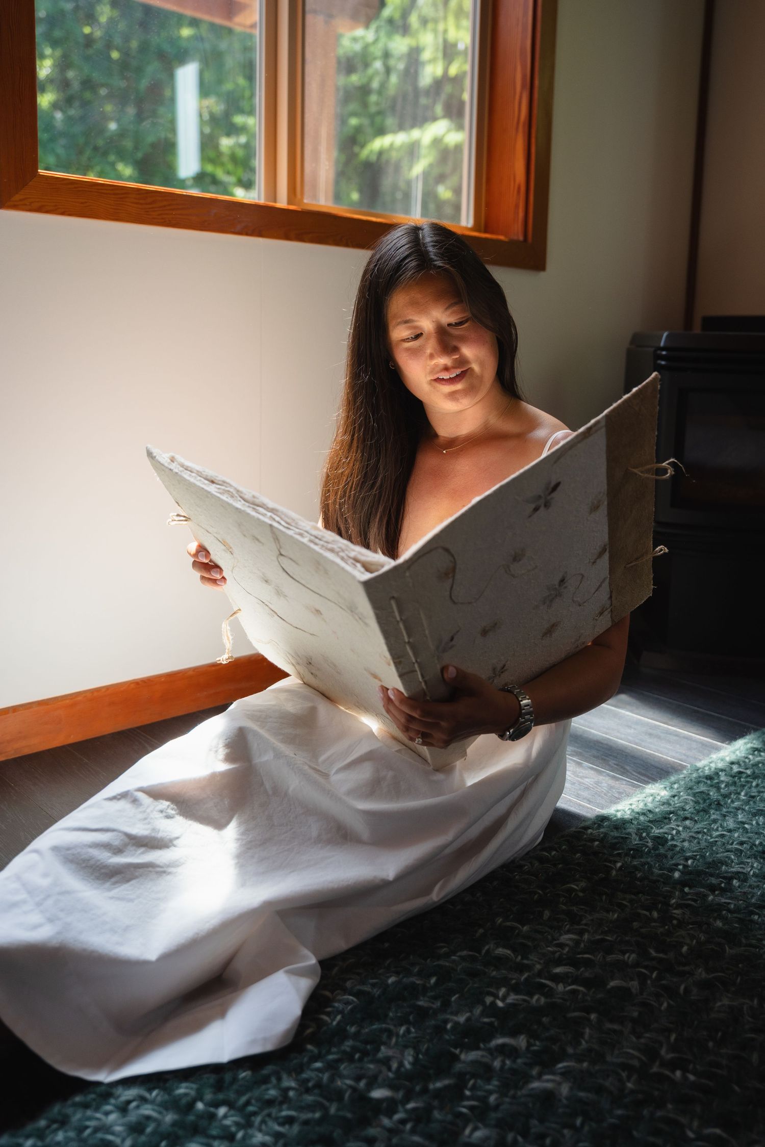 Bride in white wedding dress sitting on floor reading a vintage guest book by a sunlit window.