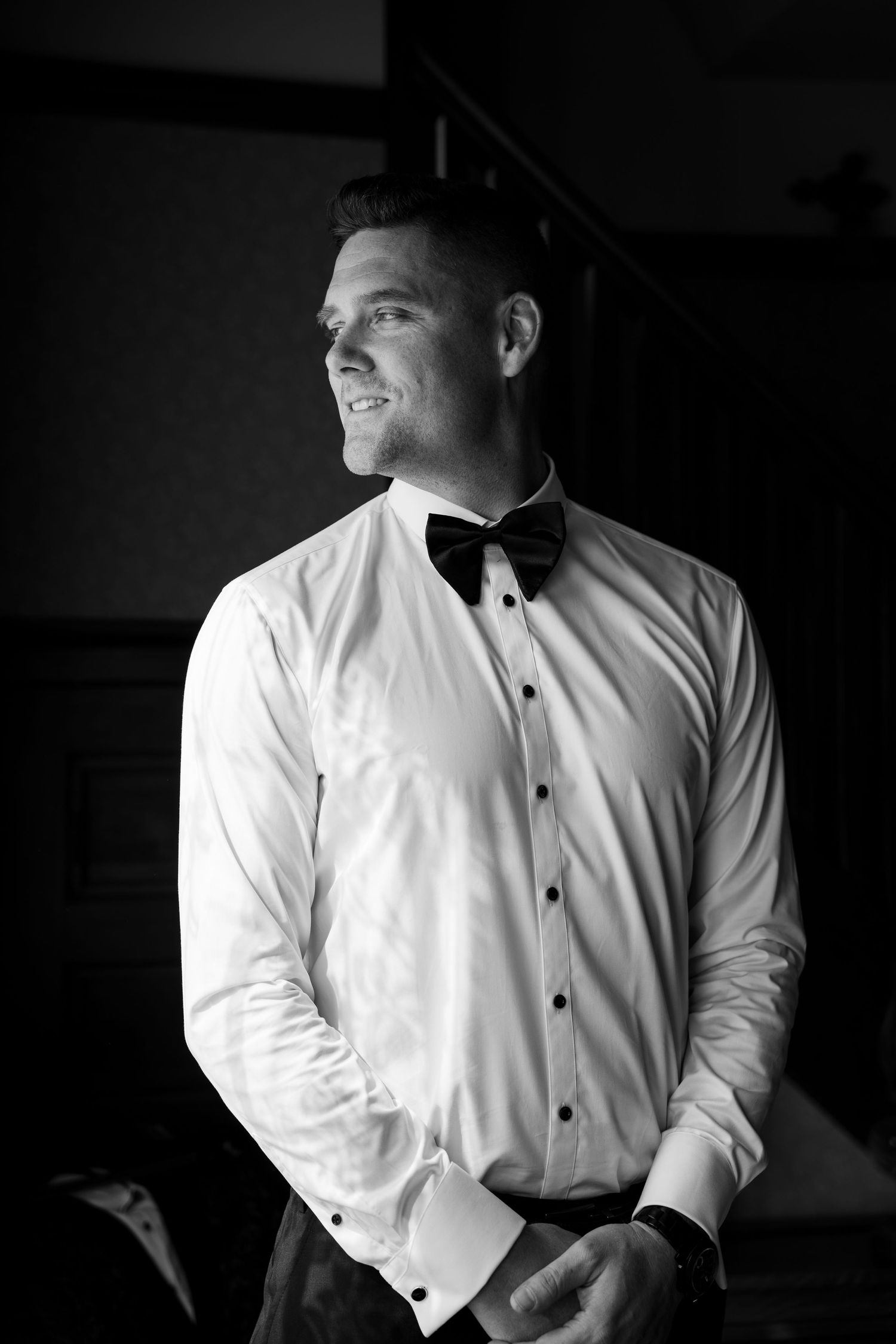 Smiling groom in white dress shirt and black bow tie looking to the side in a black and white portrait.