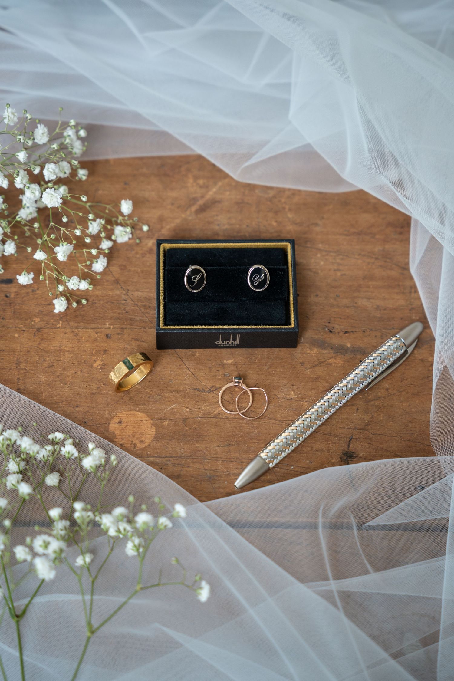 Wedding flat lay with cufflinks, gold rings, pen, and baby's breath on wood surface.
