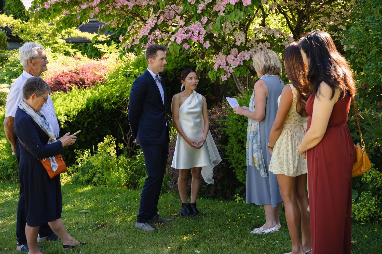 Small outdoor wedding ceremony in a lush garden with blooming pink trees, couple exchanging vows with guests.