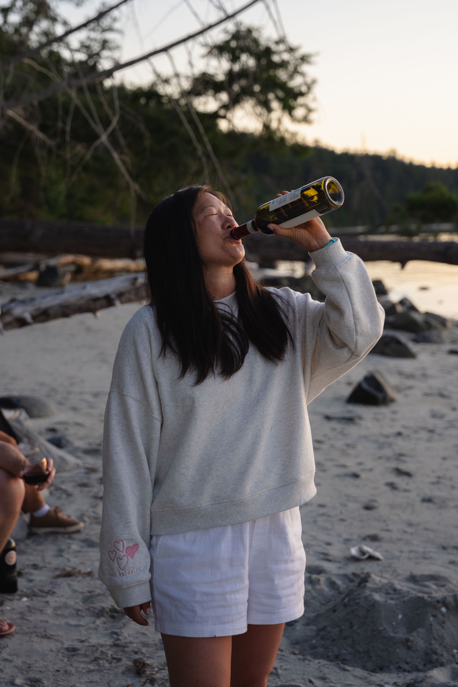 Young woman drinking from wine bottle on a rocky beach at sunset with driftwood in background.