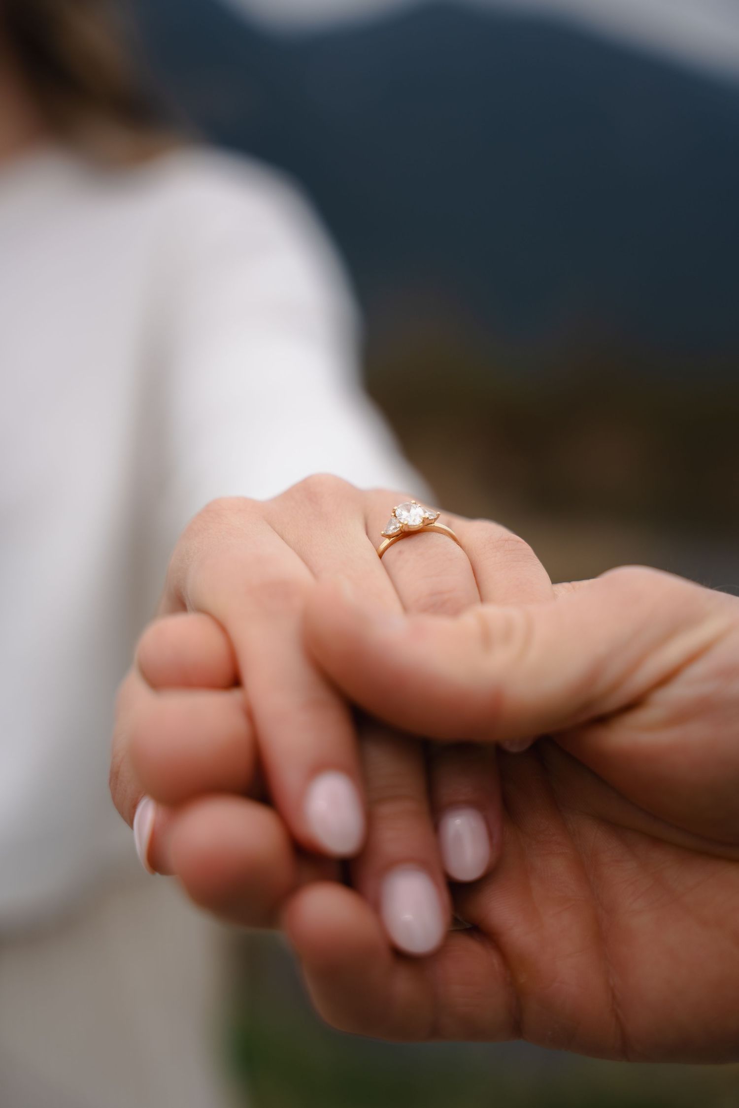 Close-up of hands holding showing a delicate gold diamond engagement ring on womans finger with mountains in background.