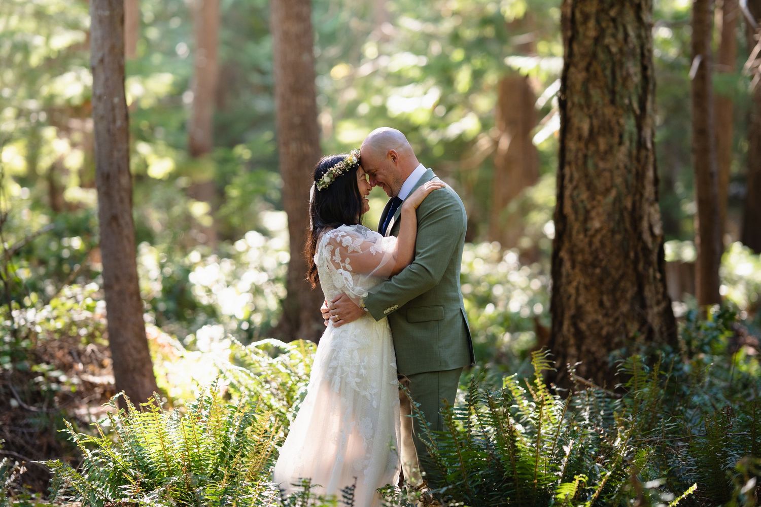Bride and groom share romantic embrace among ferns and tall trees in sunlit forest wedding photo.