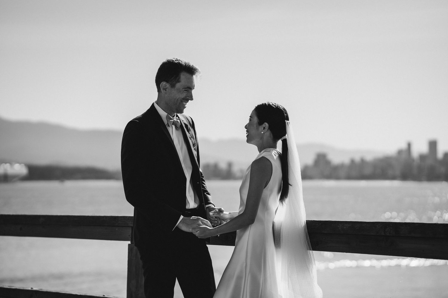 Black and white photo of bride and groom holding hands on waterfront deck with city skyline backdrop.