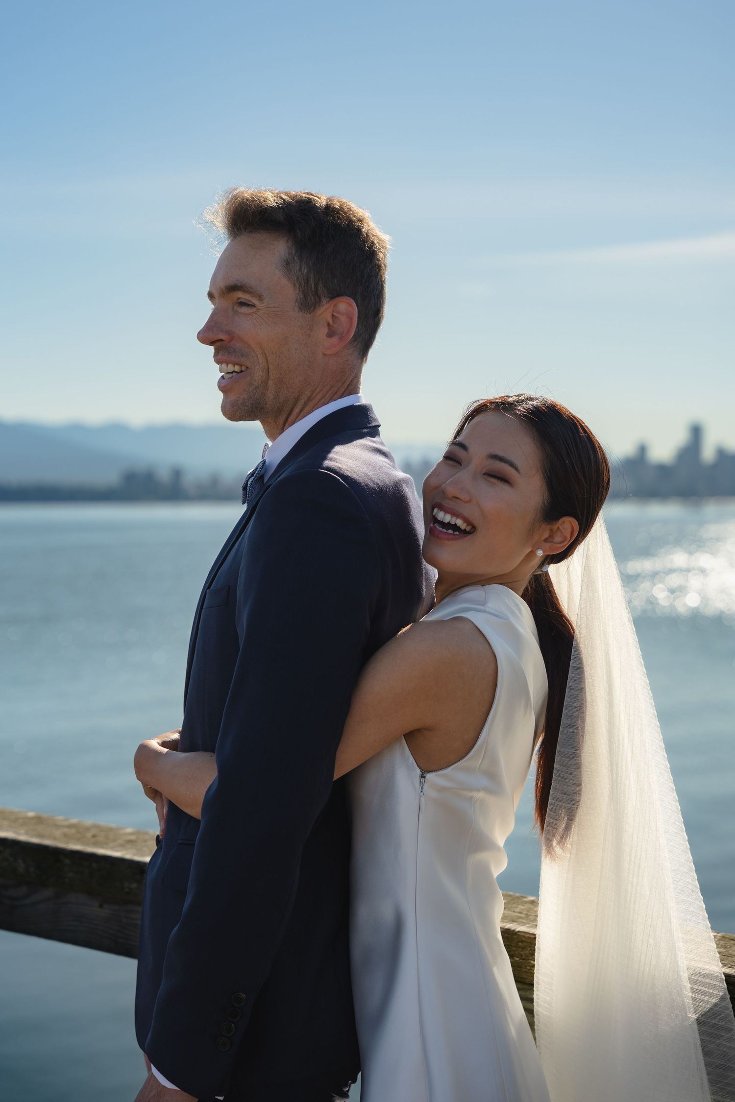Happy bride and groom laughing together by the waterfront on their wedding day, with a city skyline in the background.