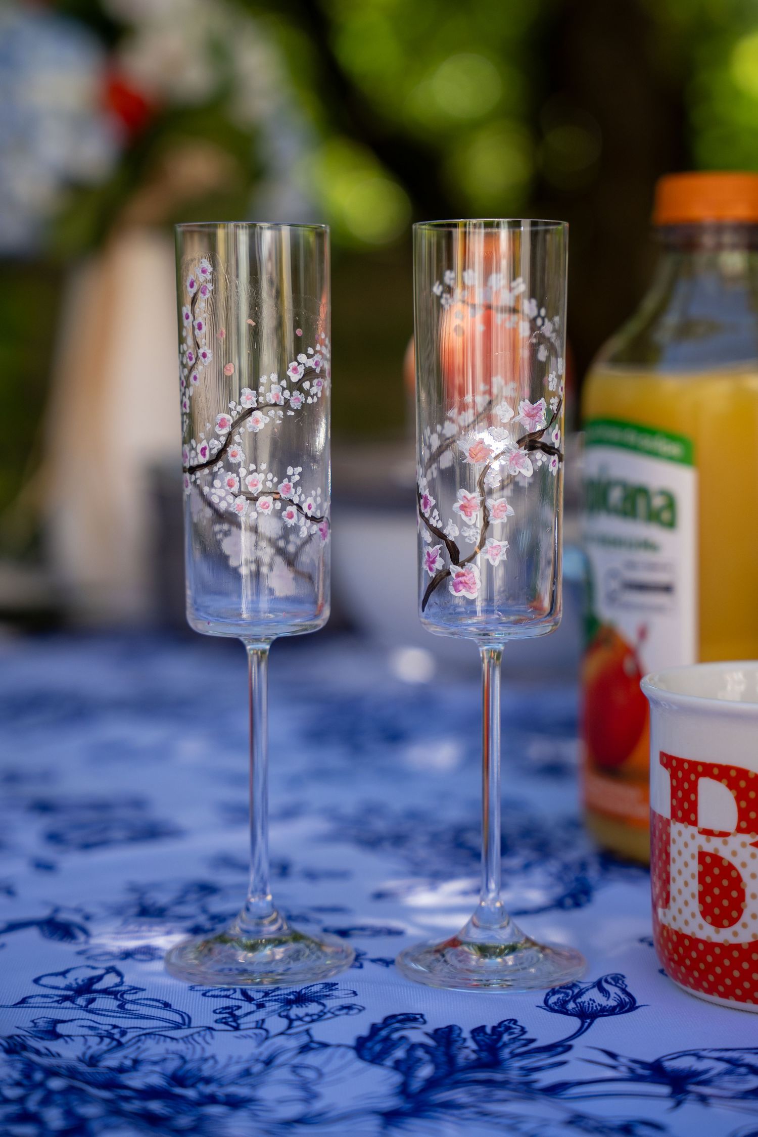 Two elegant cherry blossom painted champagne flutes on a blue floral tablecloth outdoors.
