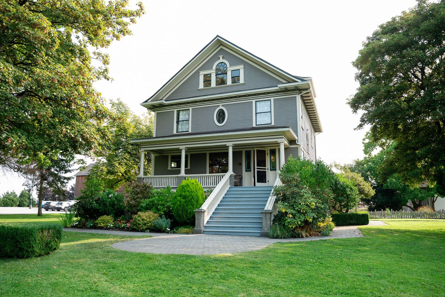 Gray two-story American Foursquare home with wrap-around porch, white trim, and lush green lawn surrounded by mature trees.