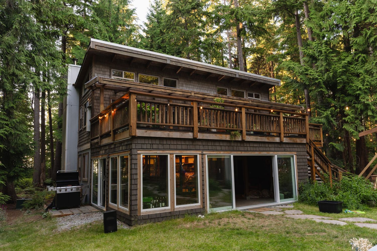 Rustic two-story wooden cabin surrounded by tall evergreen trees with large windows and deck lighting.