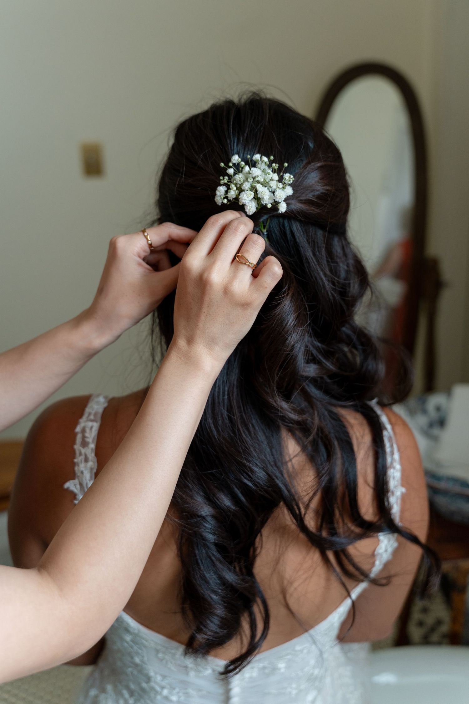 Hands placing white baby's breath flowers into bride's dark wavy hair from behind.