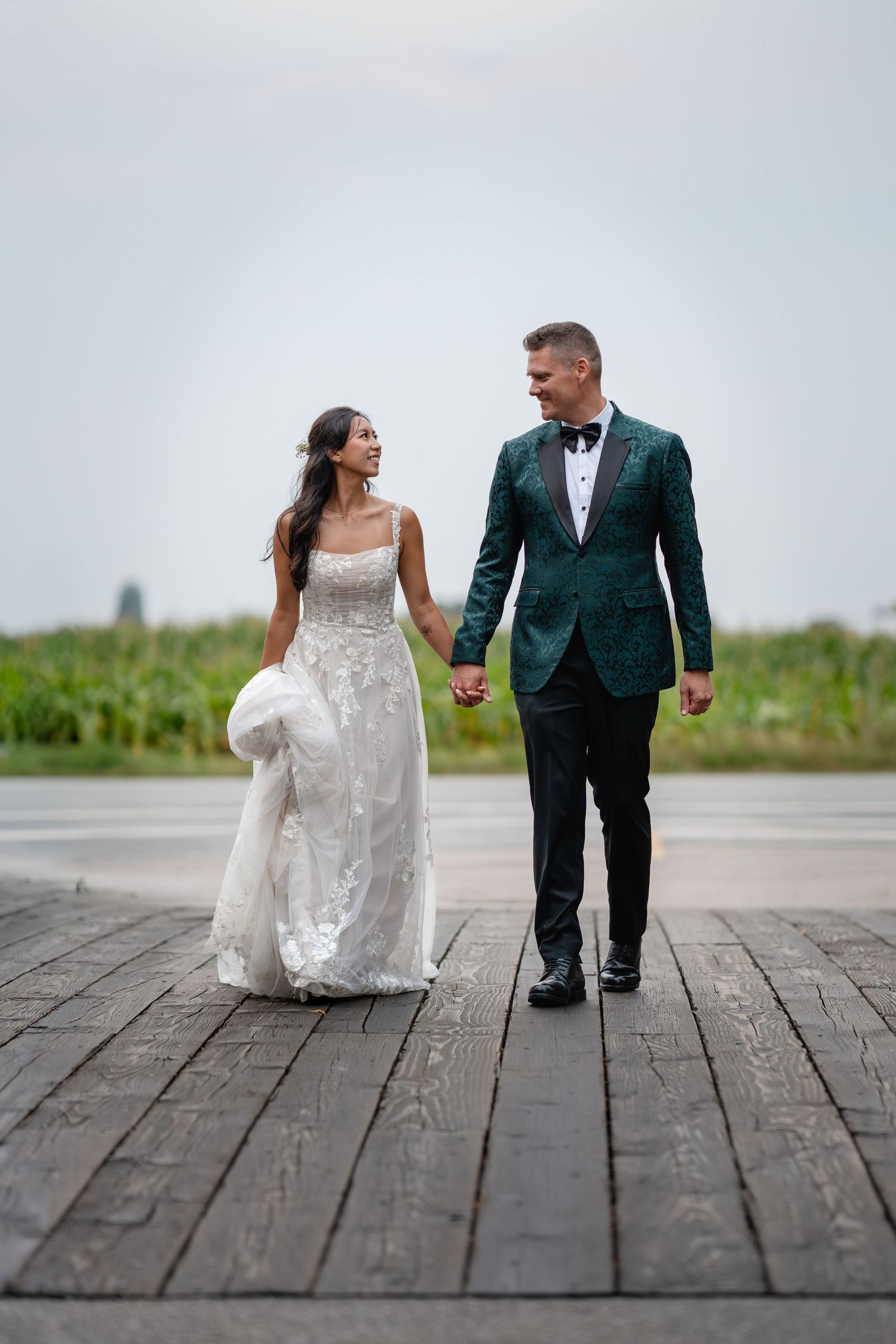 Bride and groom walk hand in hand on a wooden dock, smiling at each other on their wedding day.