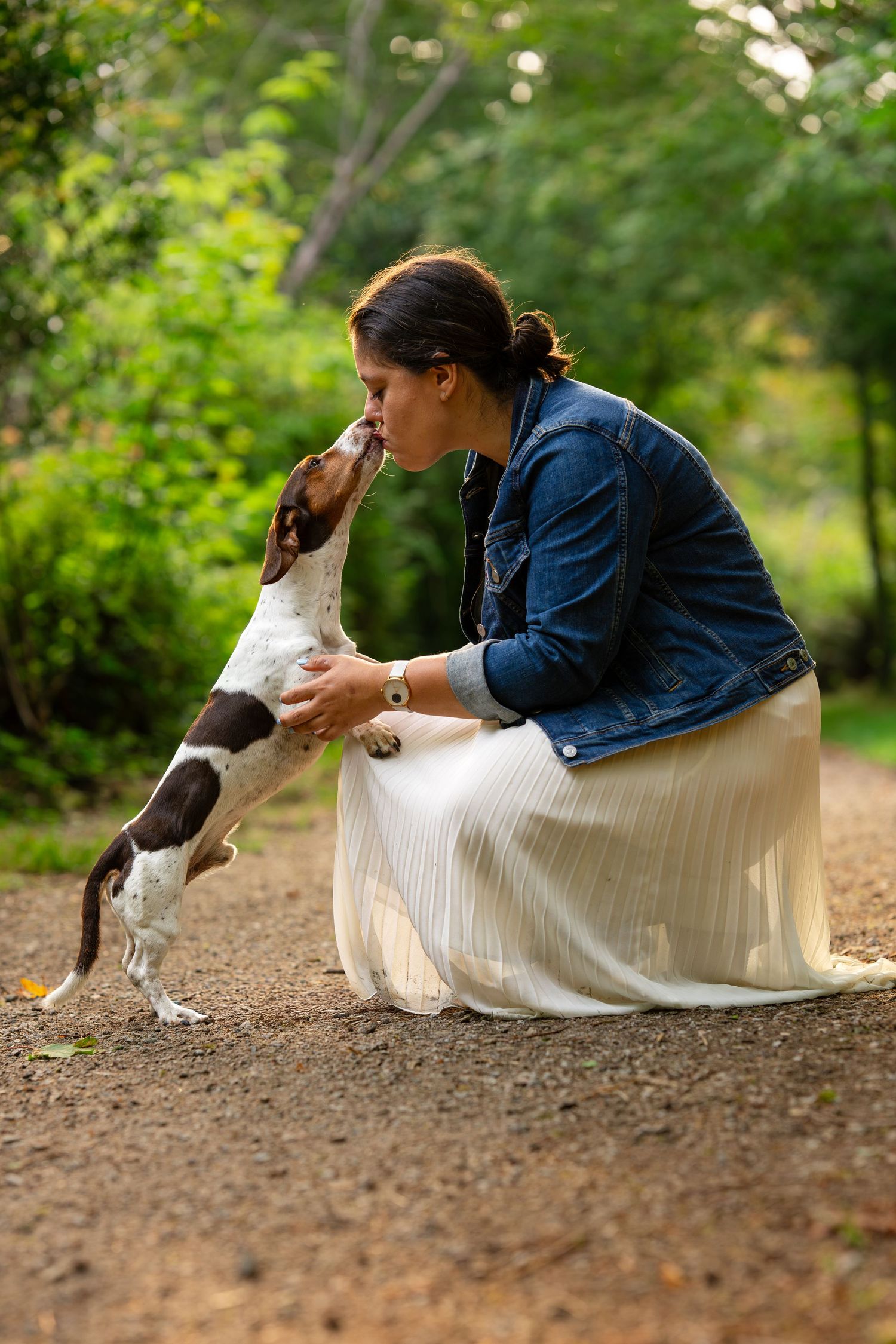 Woman in denim jacket and white skirt crouching as a beagle puppy stands up to give her a kiss outdoors.