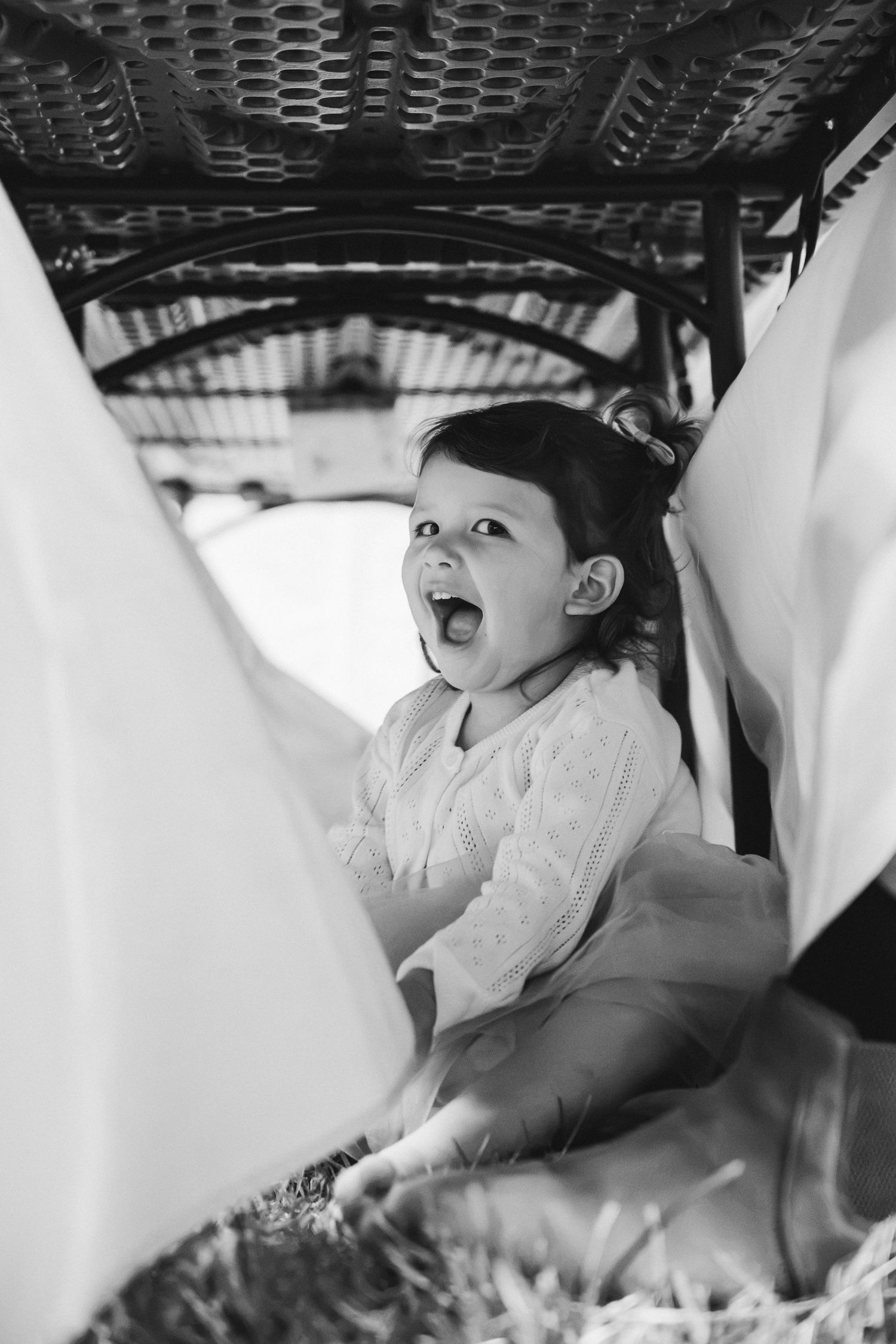 Black and white photo of a joyful toddler girl with mouth open wide sitting in a vintage wicker baby carriage.