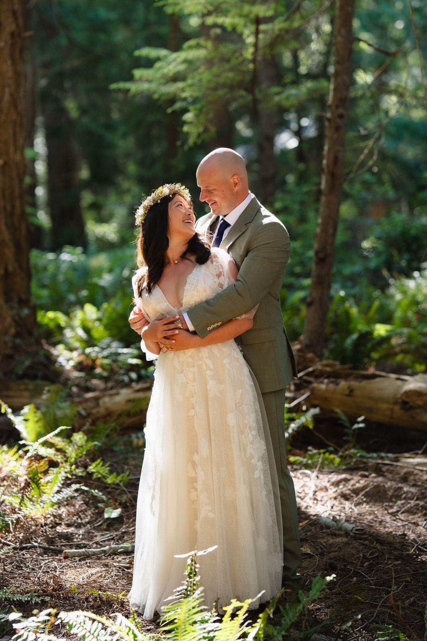 Bride in lace gown with flower crown embraced by bald groom in olive suit in sunlit forest.