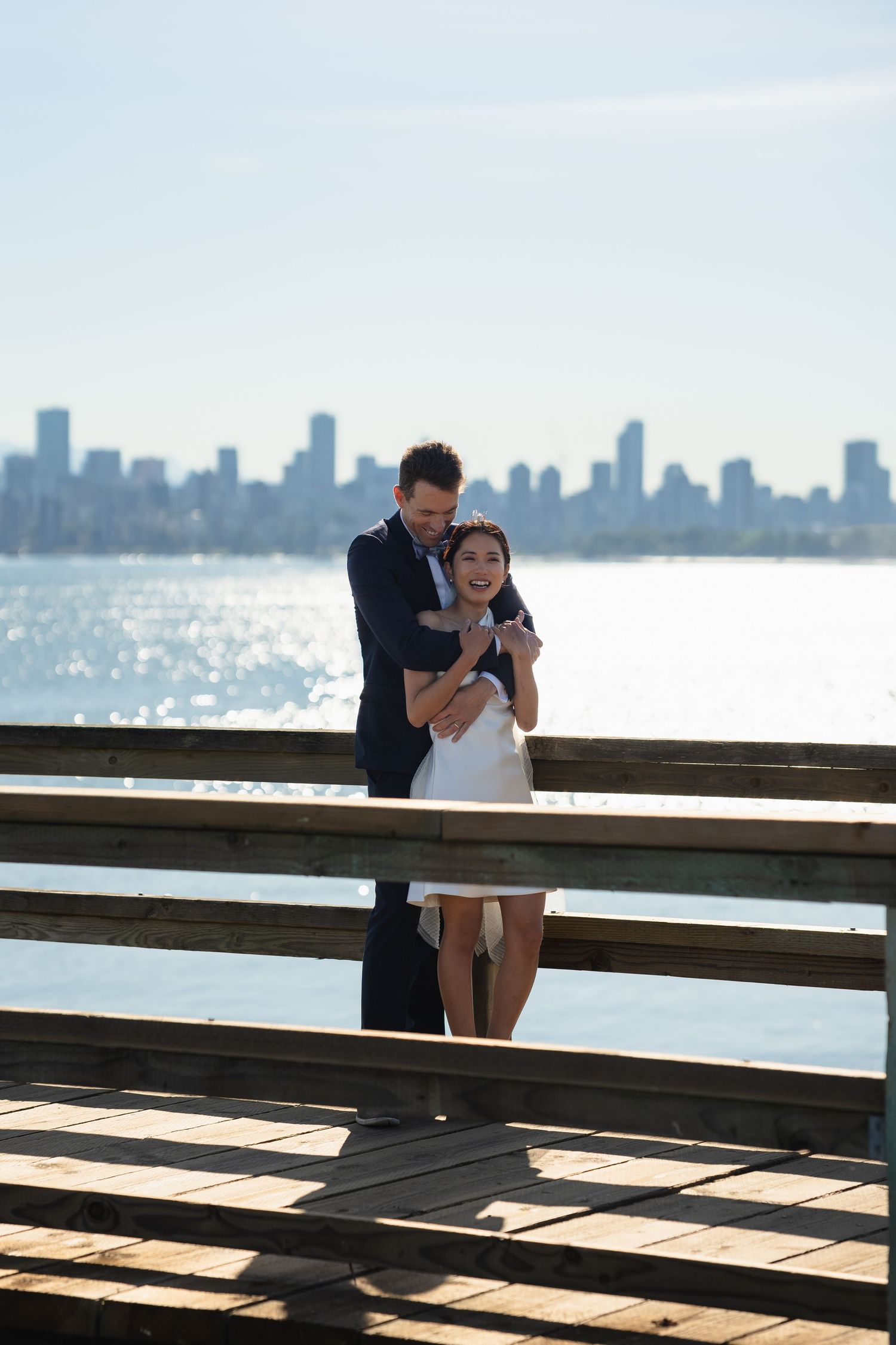 Couple embracing on a wooden pier with a city skyline and sparkling water in the background on a sunny day.