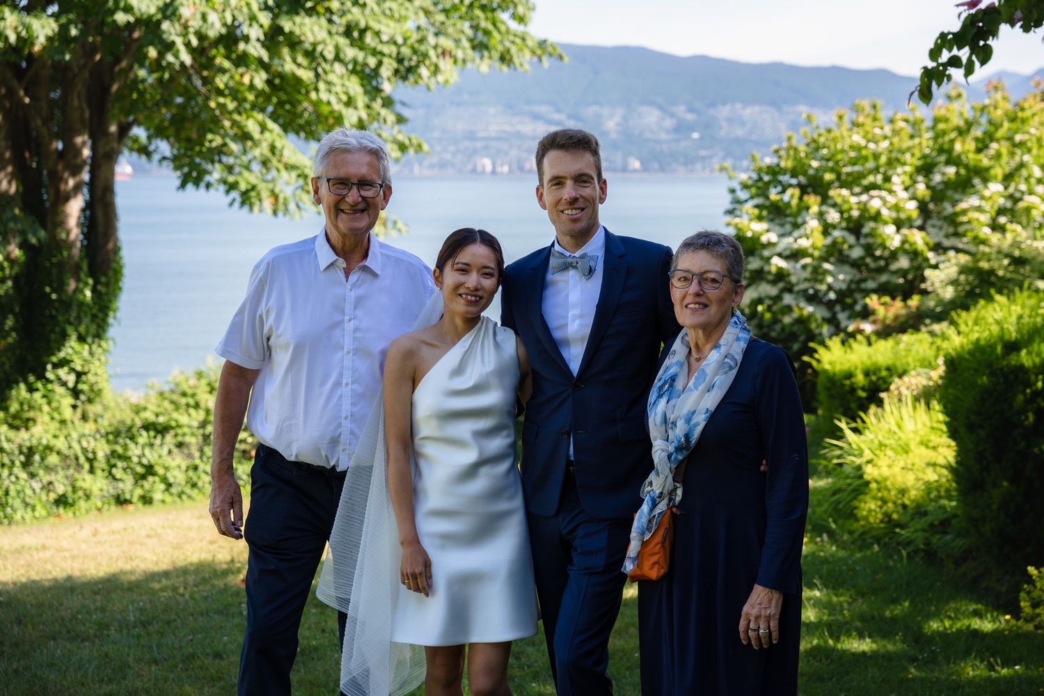 A wedding couple poses with family outdoors, with a scenic lake and hills in the background on a sunny day.