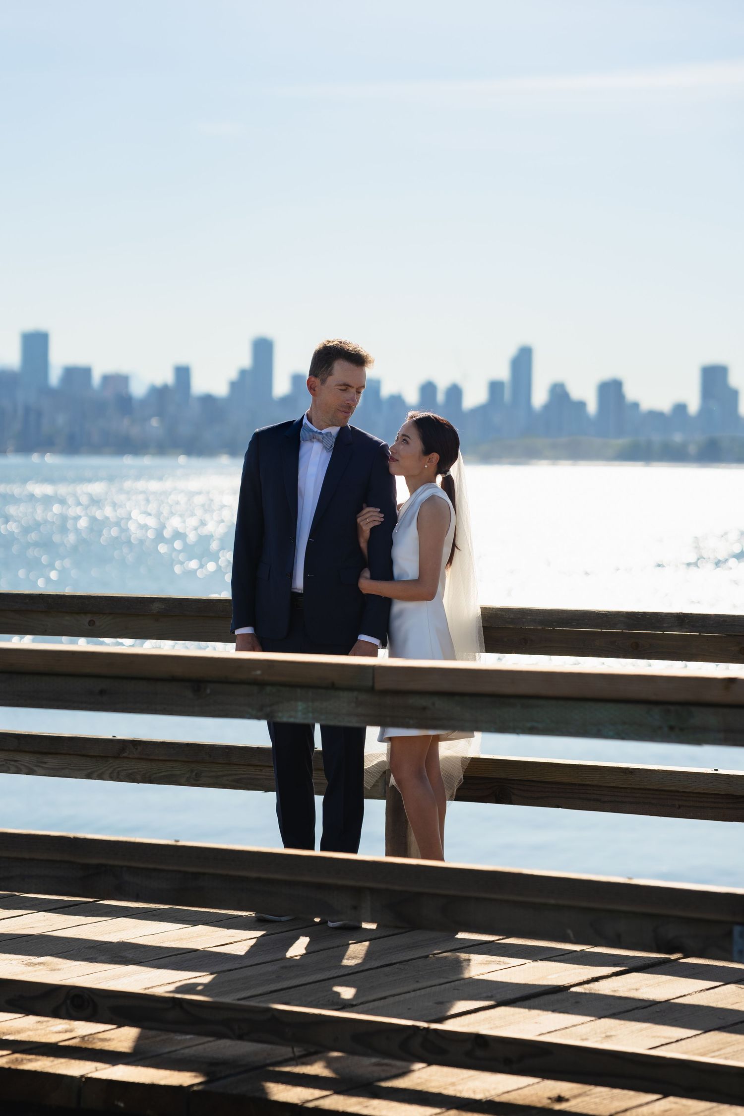 Bride and groom share a romantic moment on a wooden pier with a city skyline and sparkling water in the background.