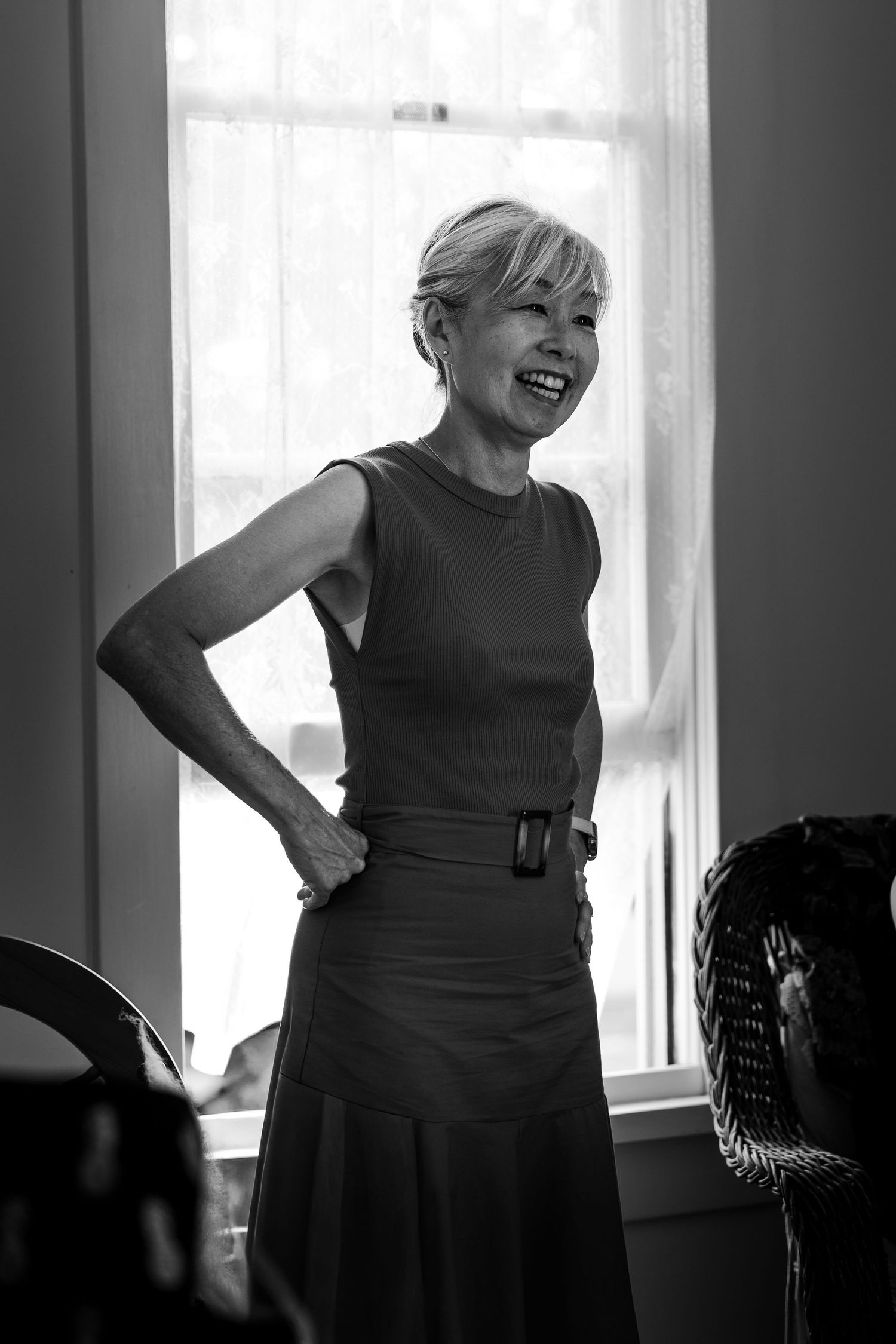 Black and white photo of a smiling older woman in a sleeveless top and skirt standing by a bright window.