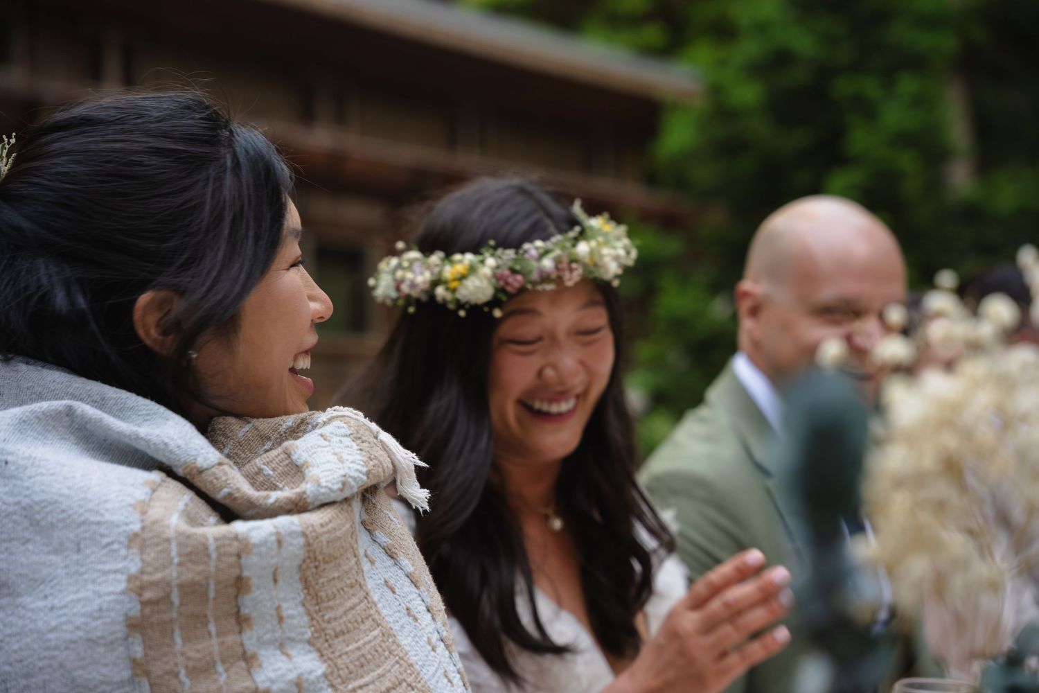 Two smiling Asian women at outdoor wedding reception, one wearing a flower crown and wrapped in a woven shawl.