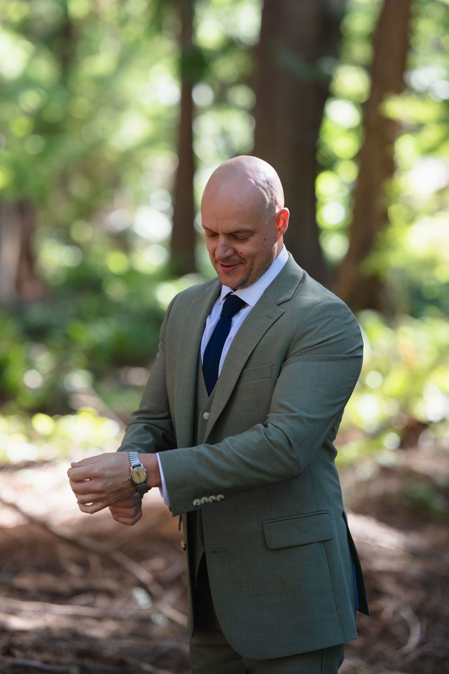 Bald groom in olive green suit checking his watch while standing in a sunlit forest.