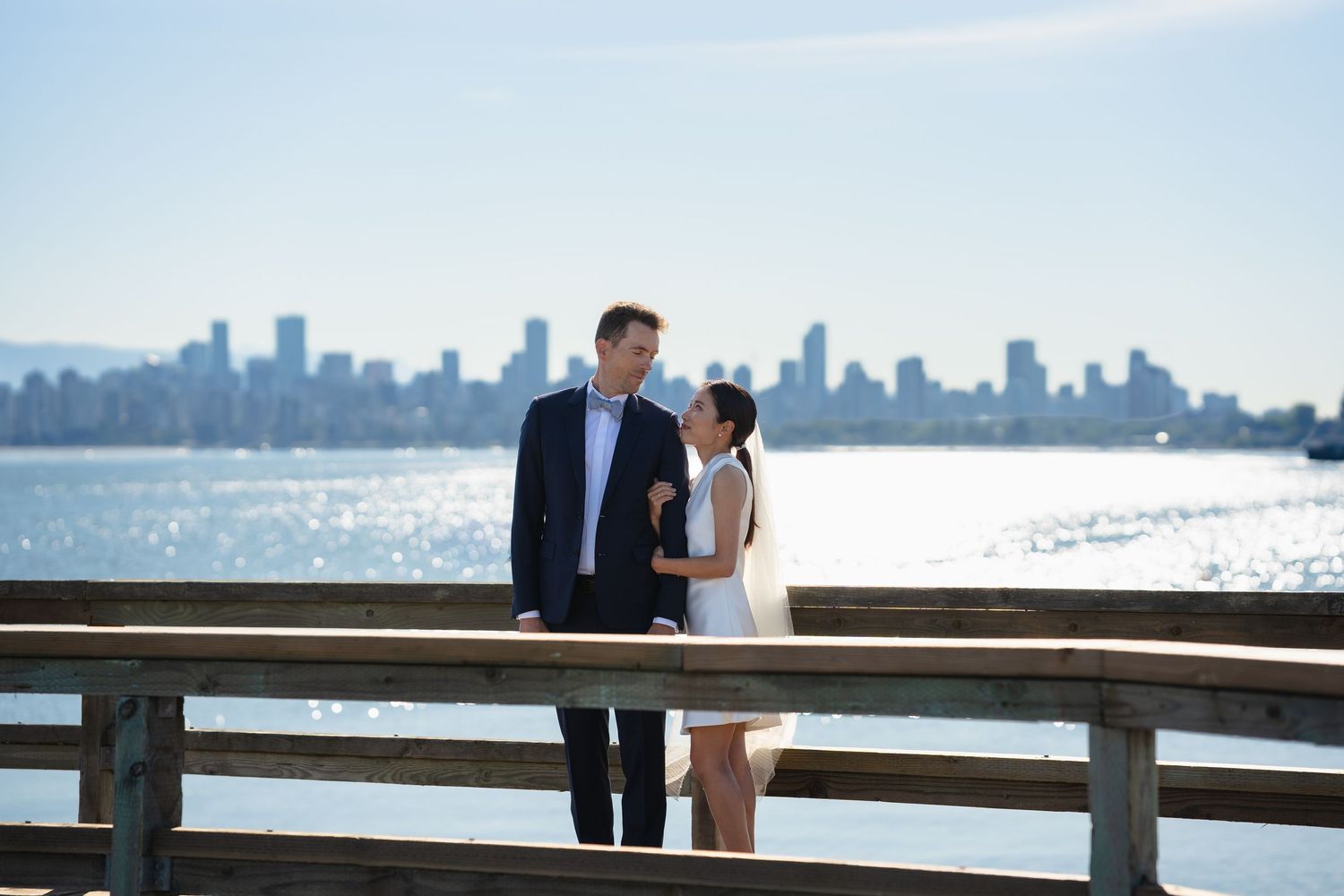 Couple shares a romantic moment on a wooden pier with a stunning city skyline and calm blue water in the background.