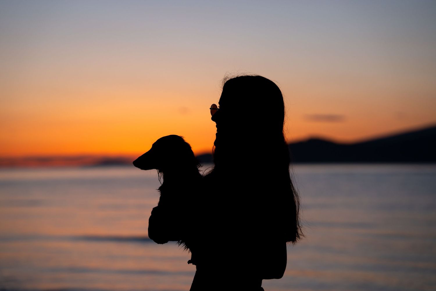 Silhouette of a woman holding a small dog against a vibrant orange and pink sunset over the ocean.