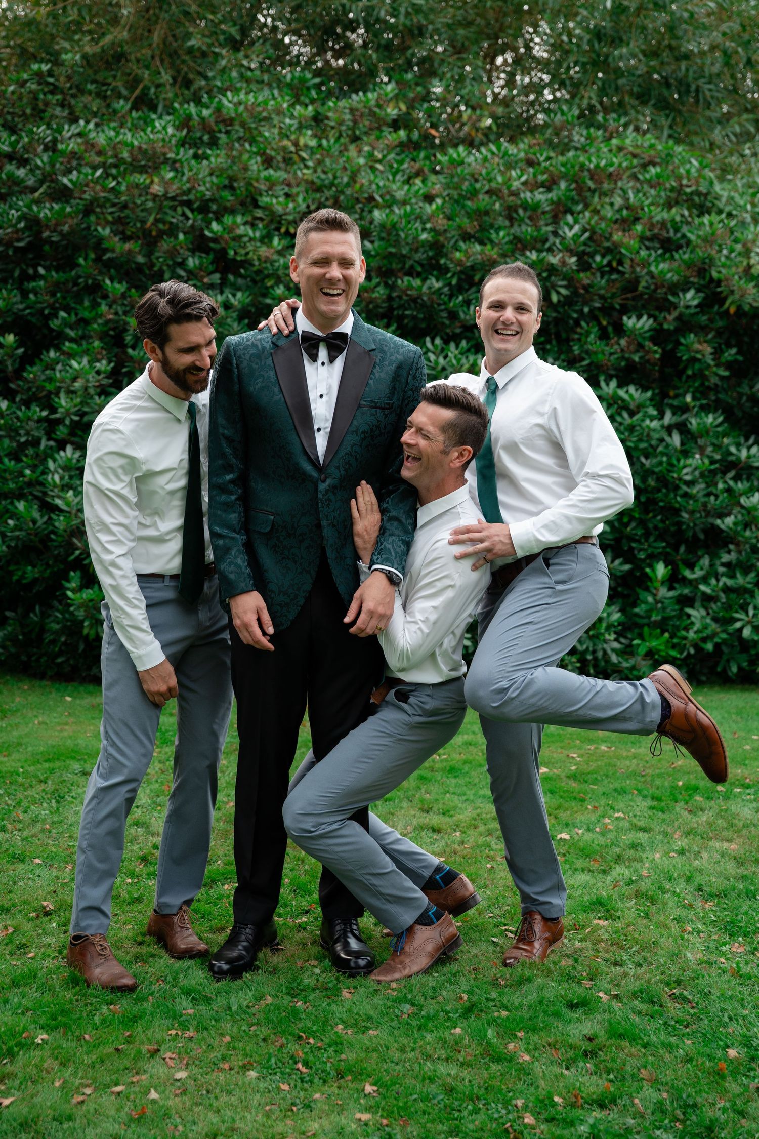Four groomsmen laughing and posing together outdoors in suits on green grass with lush hedges behind them.