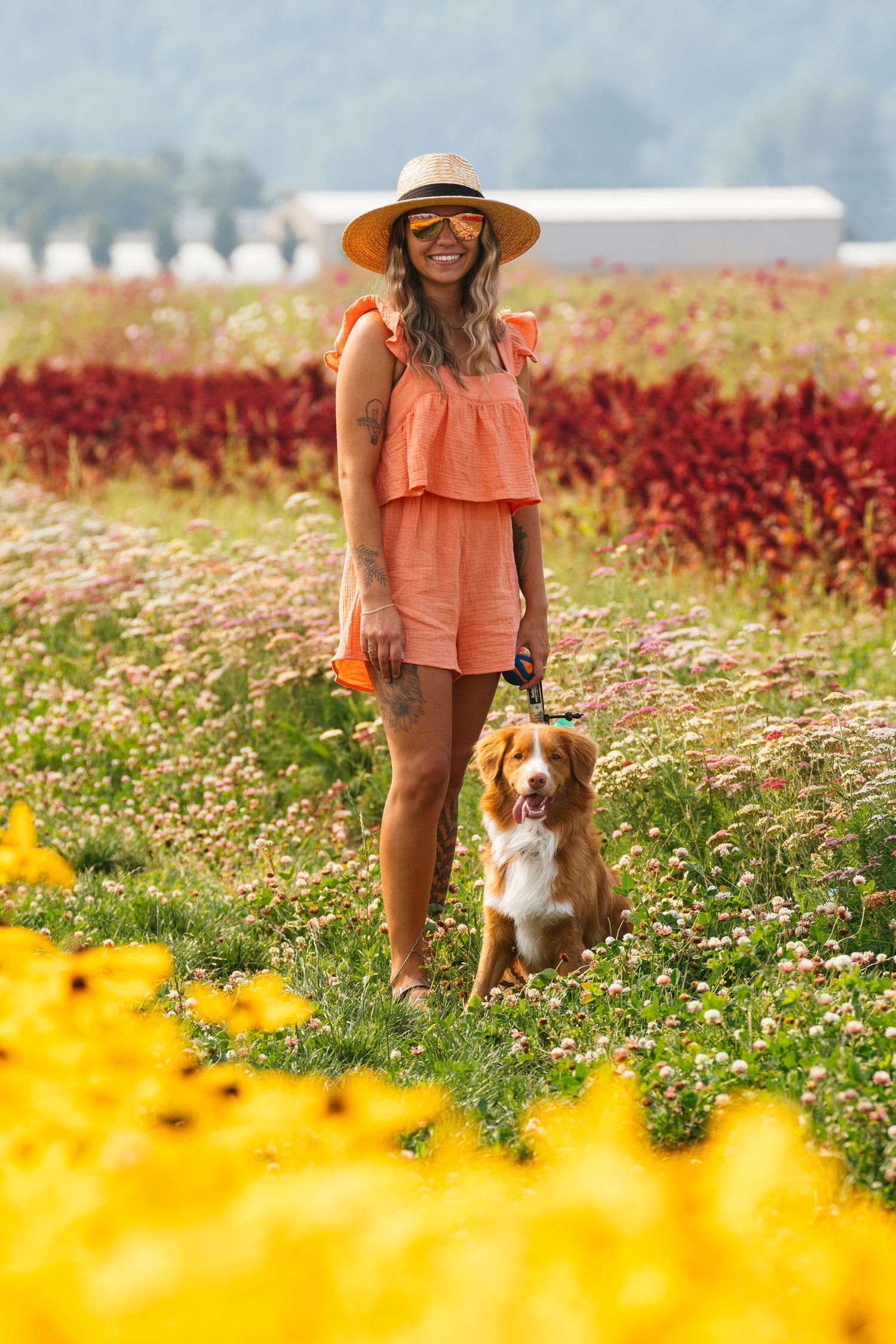 Smiling woman in orange romper and sun hat standing with Nova Scotia Duck Tolling Retriever in colorful flower field.