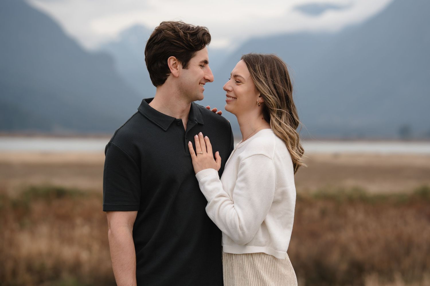 Couple sharing a romantic moment outdoors with mountain backdrop, woman in white dress touching man's chest.