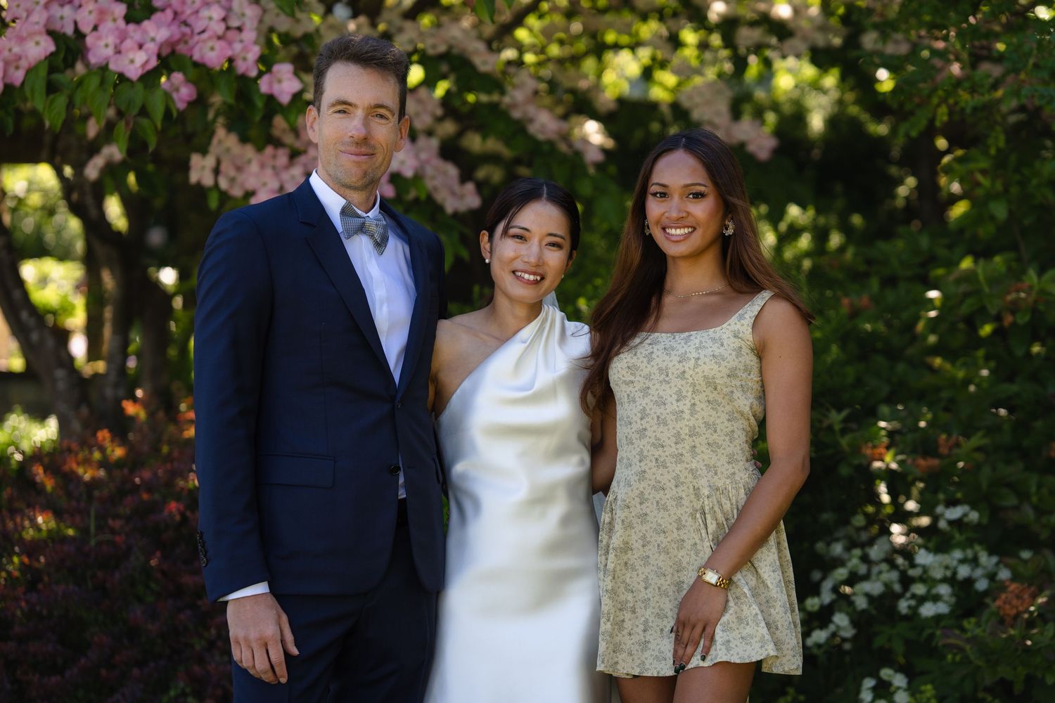 Bride in white dress poses with groom in navy suit and female guest under blooming pink trees.