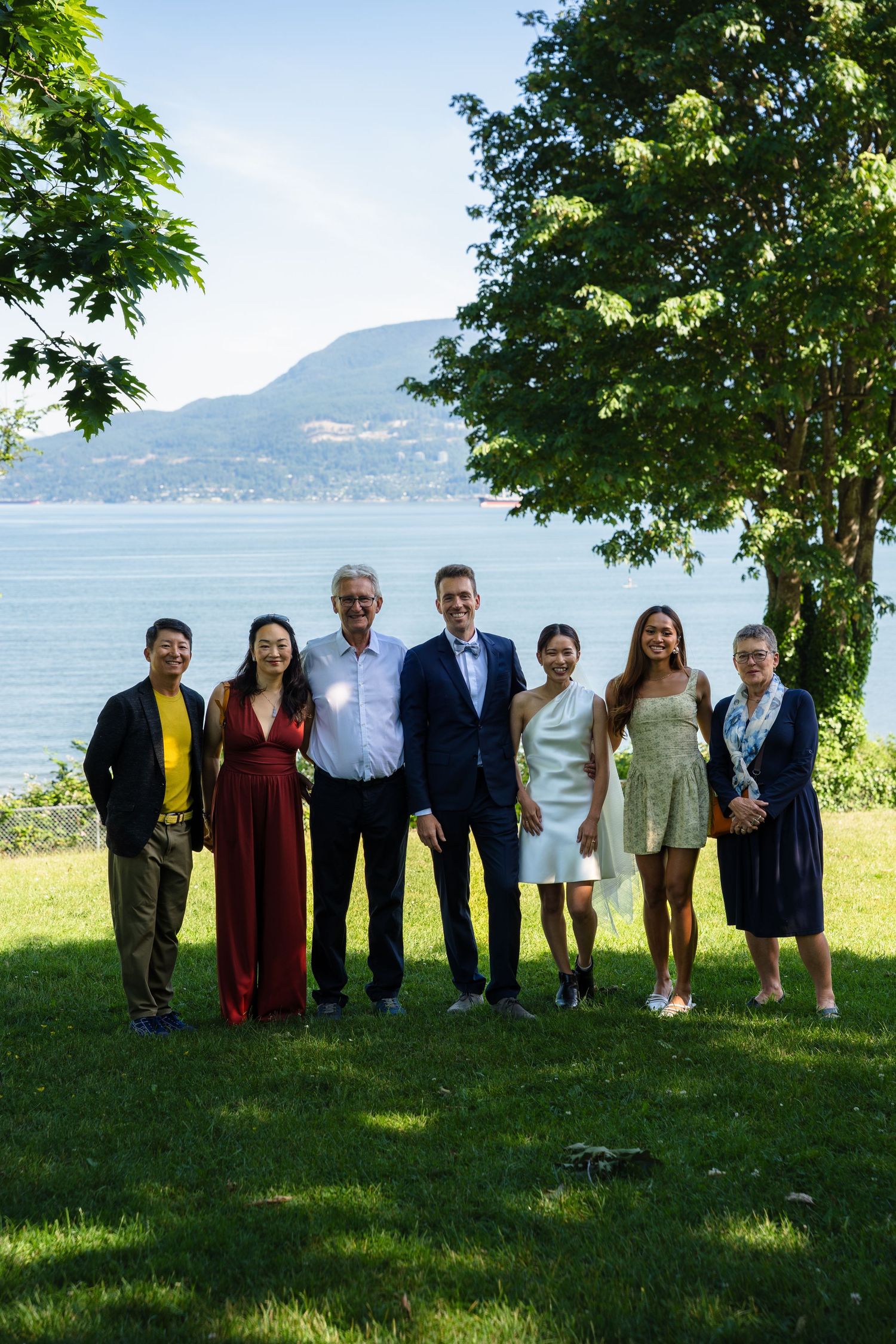 Group of seven people posing outdoors by a lake with mountain views on a sunny day.