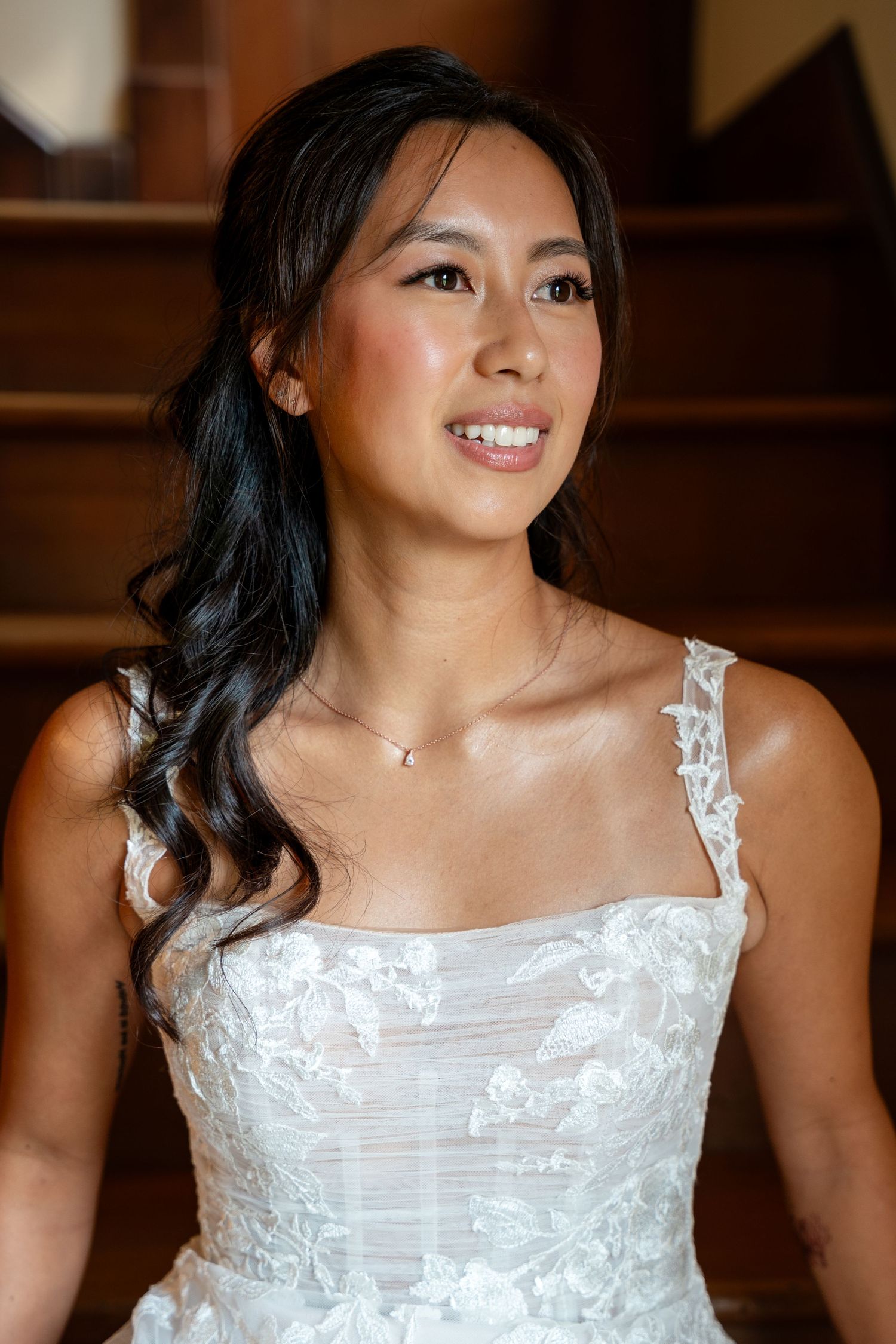 A smiling bride in a white lace wedding dress with elegant half-up hairstyle sitting on stairs.
