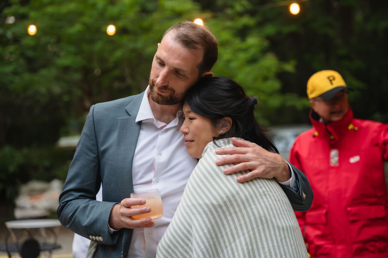 A bearded man in a gray blazer hugs a woman in a striped wrap at an outdoor evening gathering with string lights.