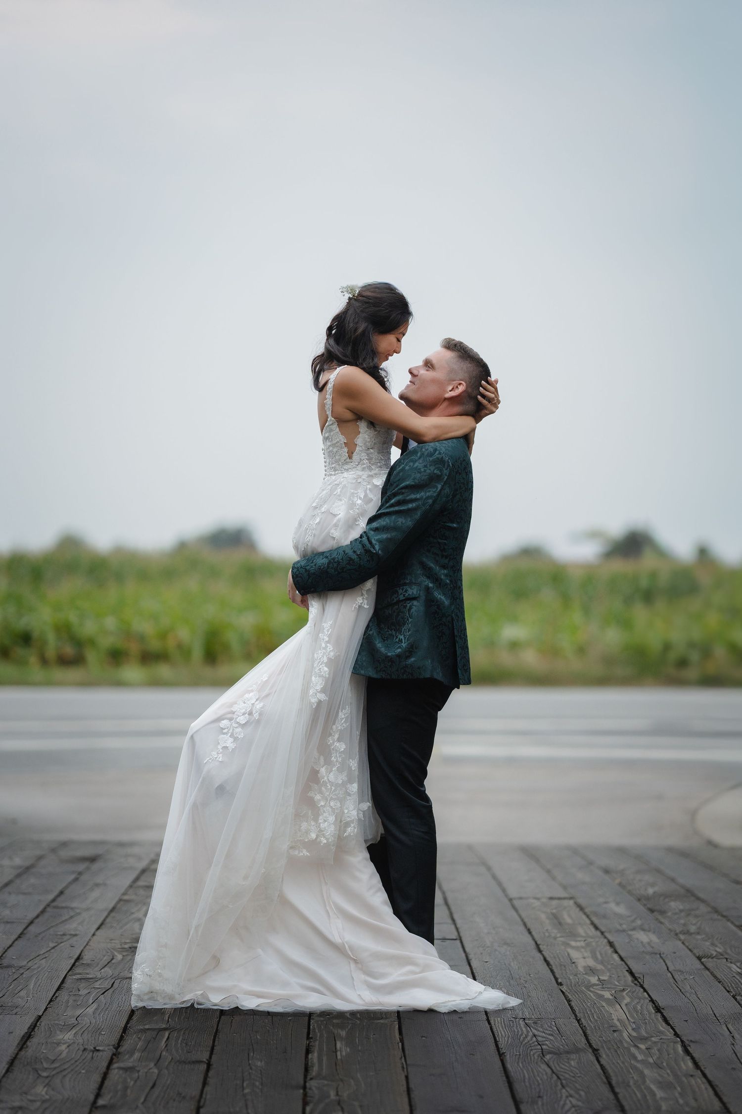 A groom lifts his bride in a white lace wedding dress on a wooden dock with greenery behind them.