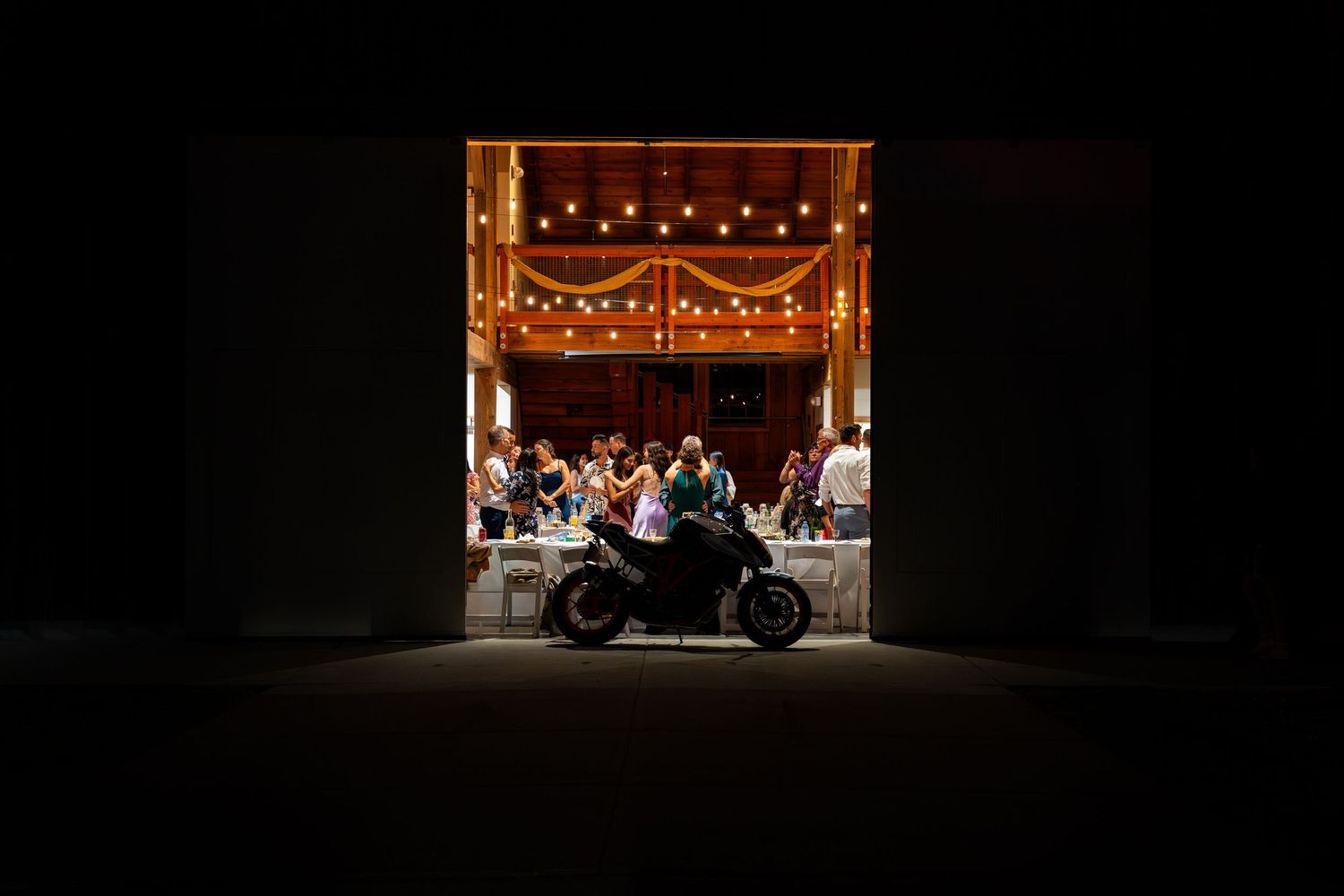 Silhouette of motorcycle framing warmly lit wedding reception in rustic barn.