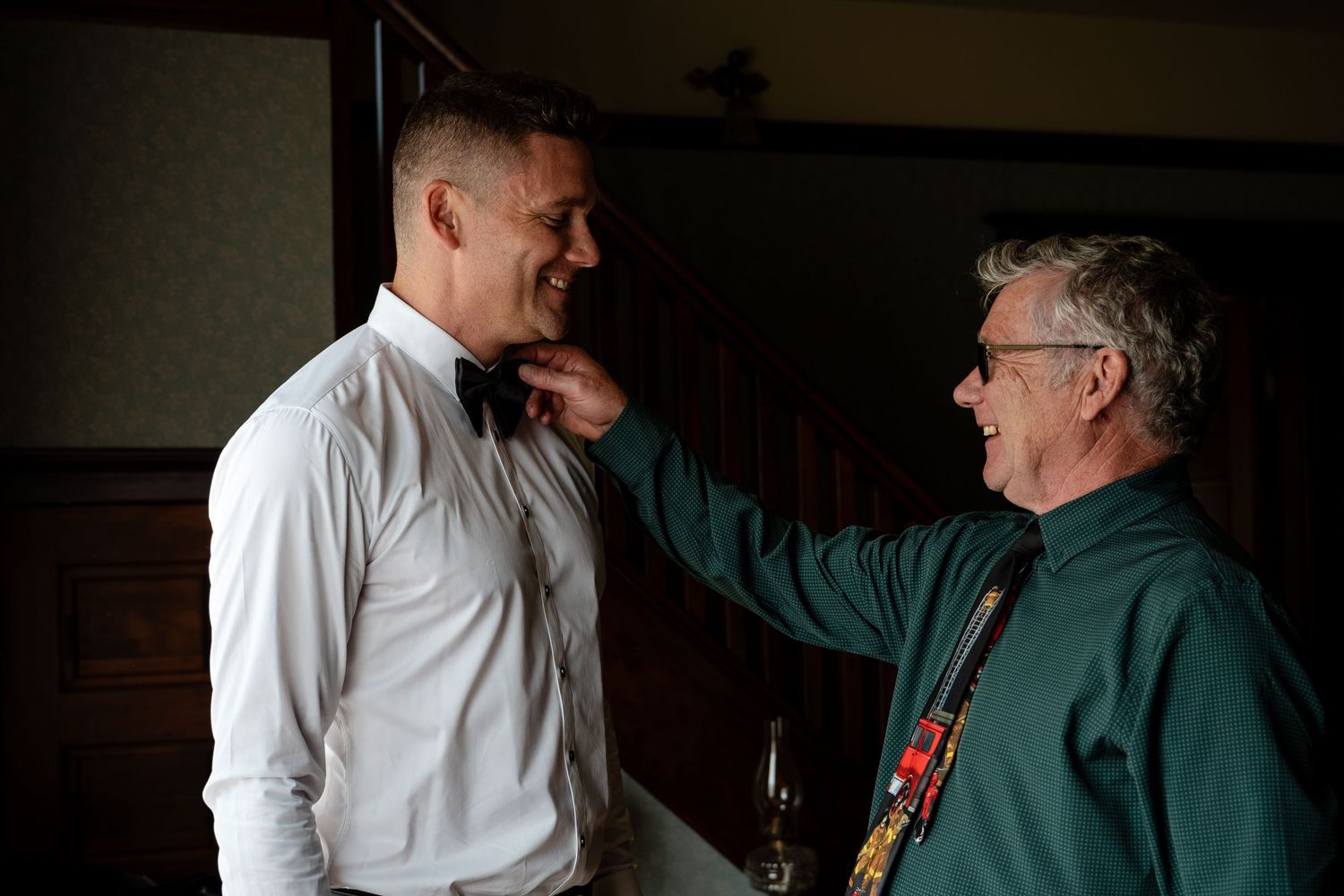 An older man adjusts a bow tie on a younger man in formal attire during wedding preparations indoors.