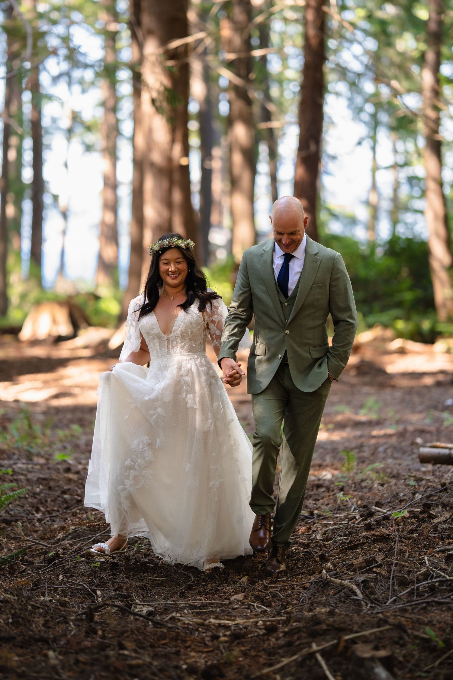 Happy bride in floral crown and groom in green suit walk hand in hand through a forest.