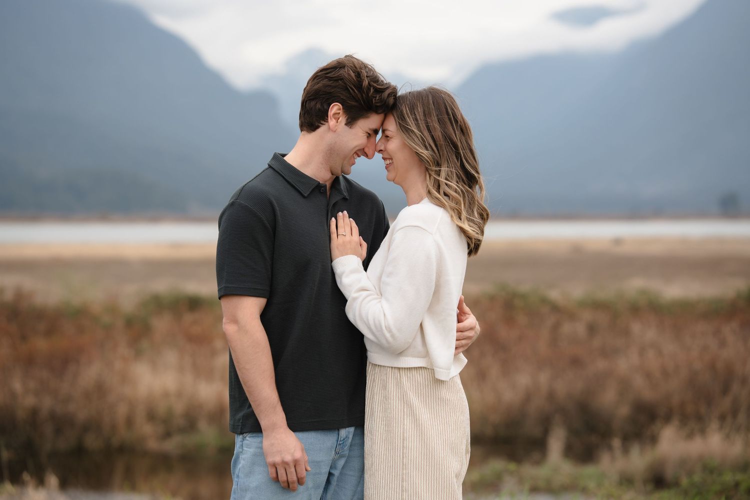 A couple shares a romantic moment outdoors, touching foreheads with mountains and a misty lake in the background.