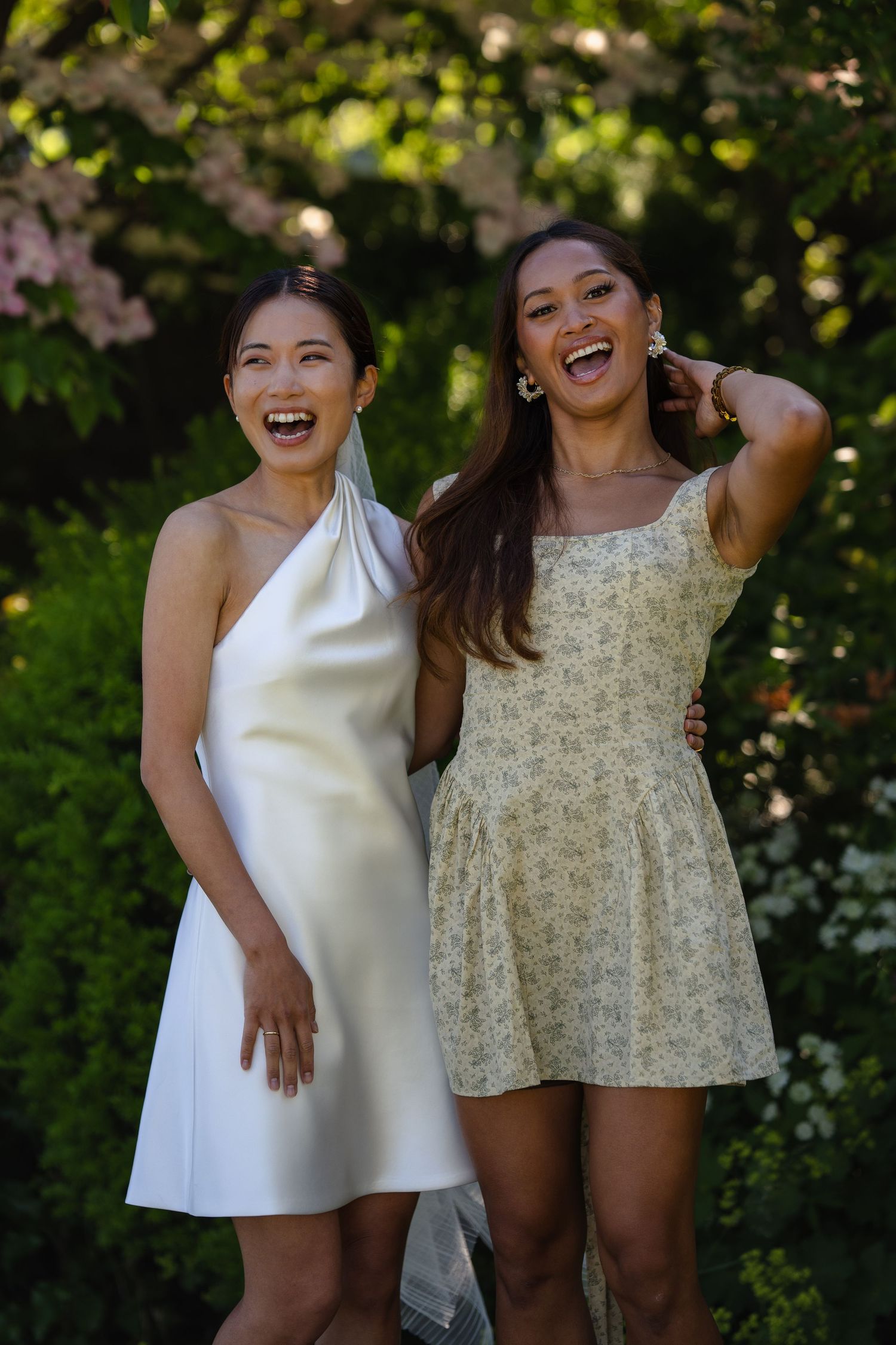 Two smiling women in elegant mini dresses pose together in a lush garden with blooming flowers.