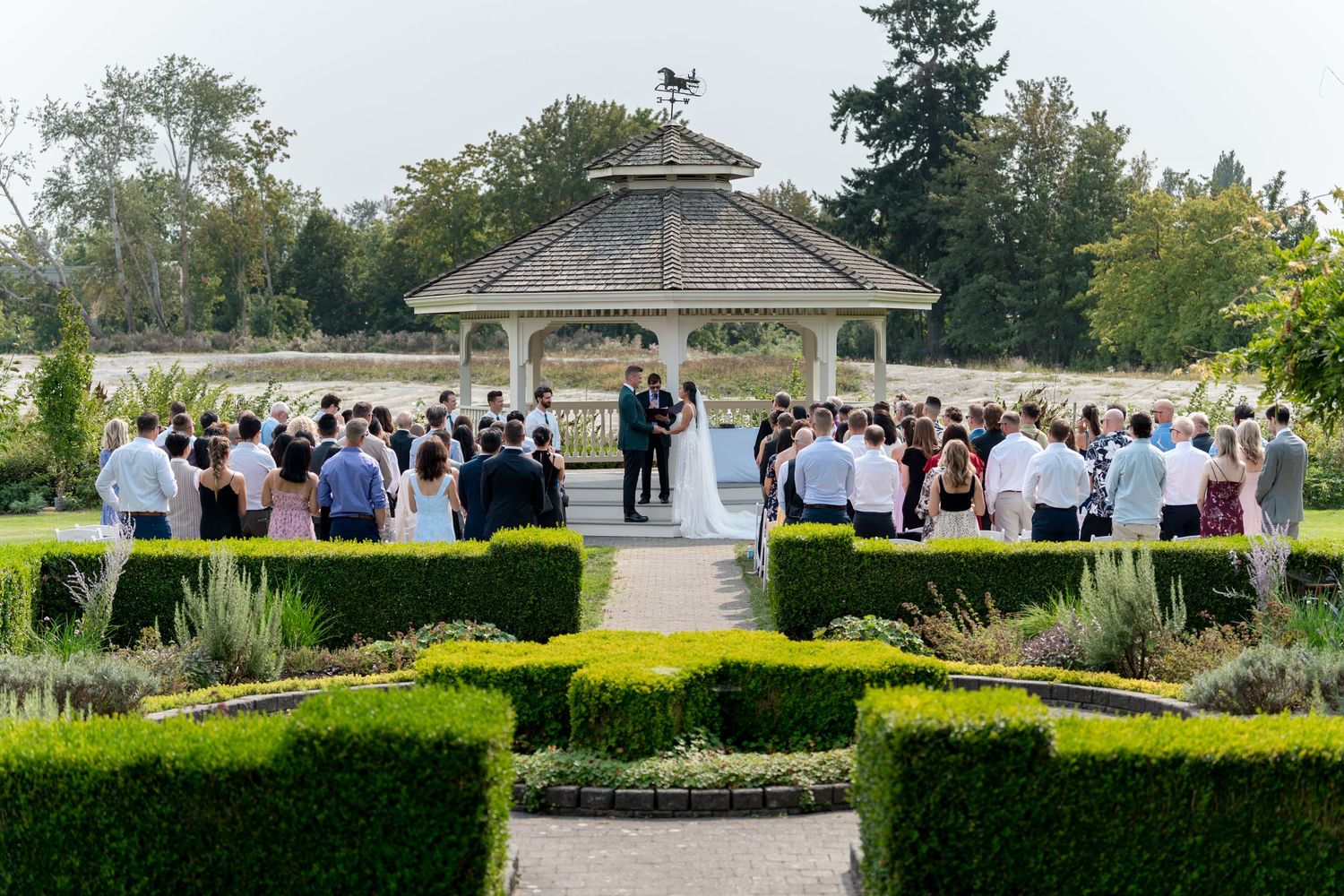 Outdoor wedding ceremony at a gazebo surrounded by manicured hedges and guests in a lush garden setting.