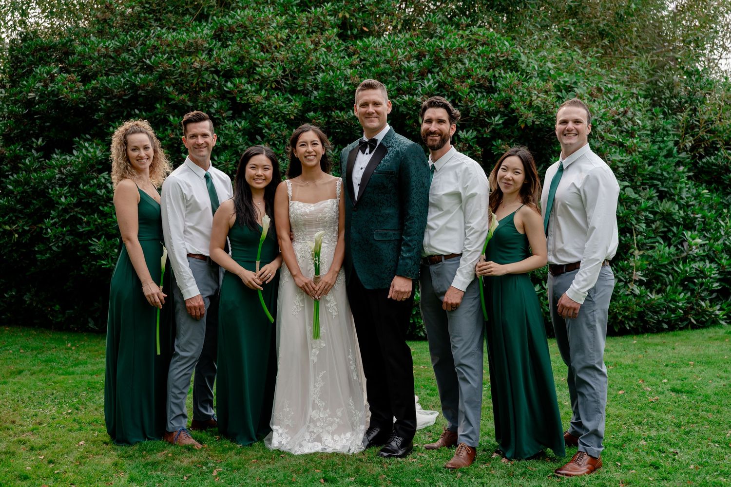 Wedding party posing together in green bridesmaid dresses and suits with bride and groom centered outdoors.
