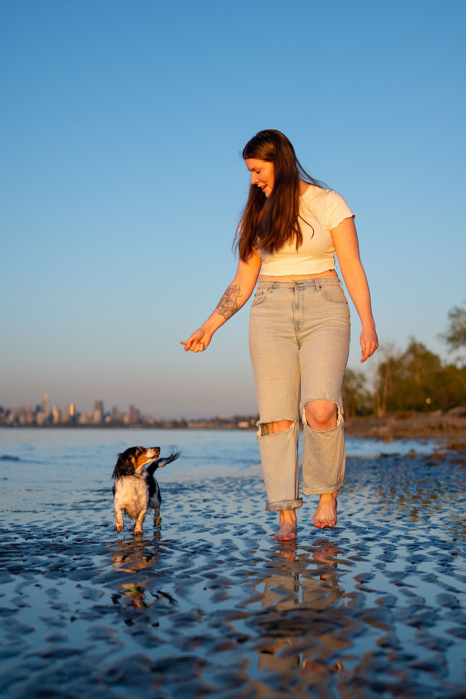 Woman in jeans and white crop top walks barefoot with small dog in shallow water at sunset on a beach.