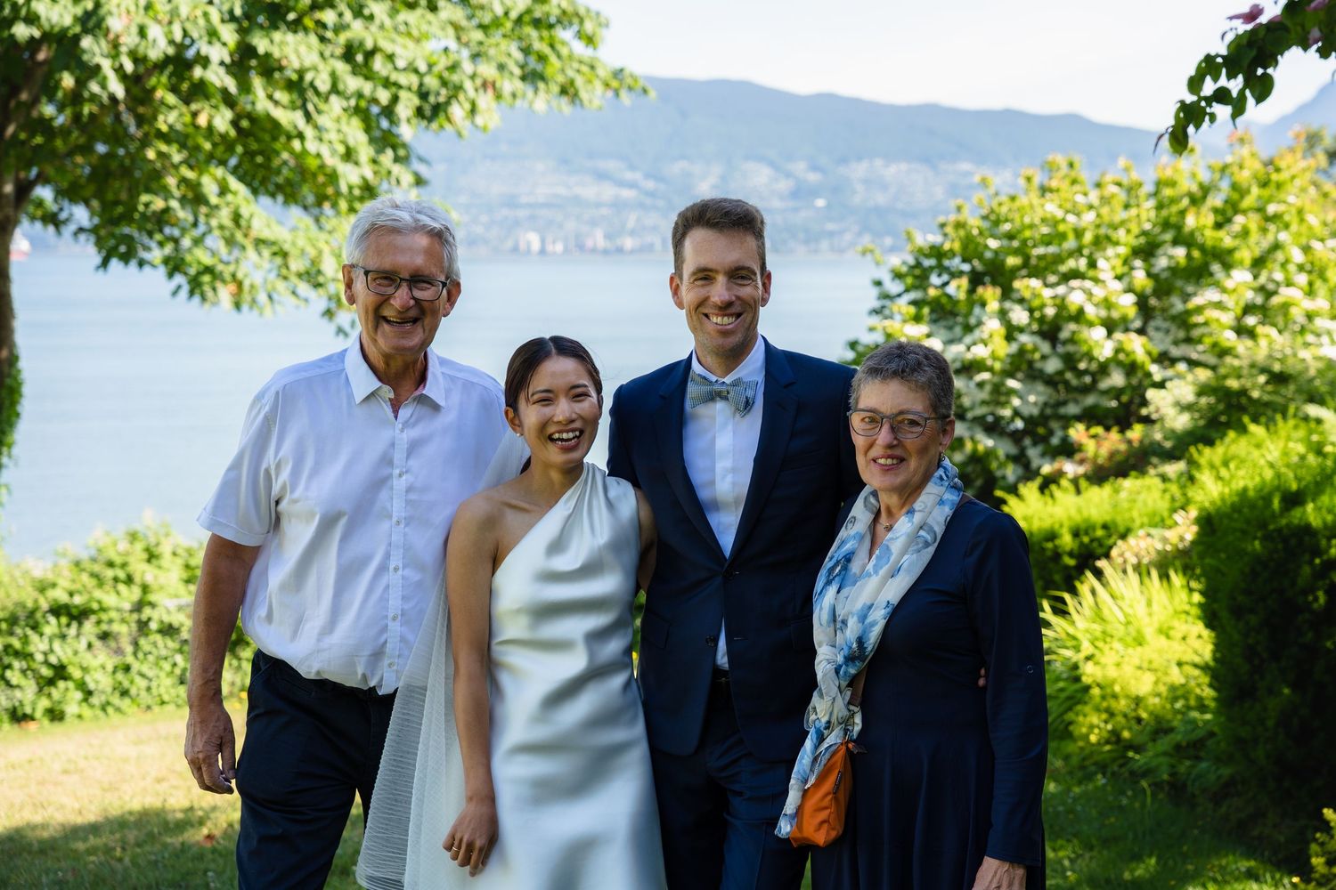 Family of four posing outdoors at a wedding celebration with a scenic lake and green trees in the background.