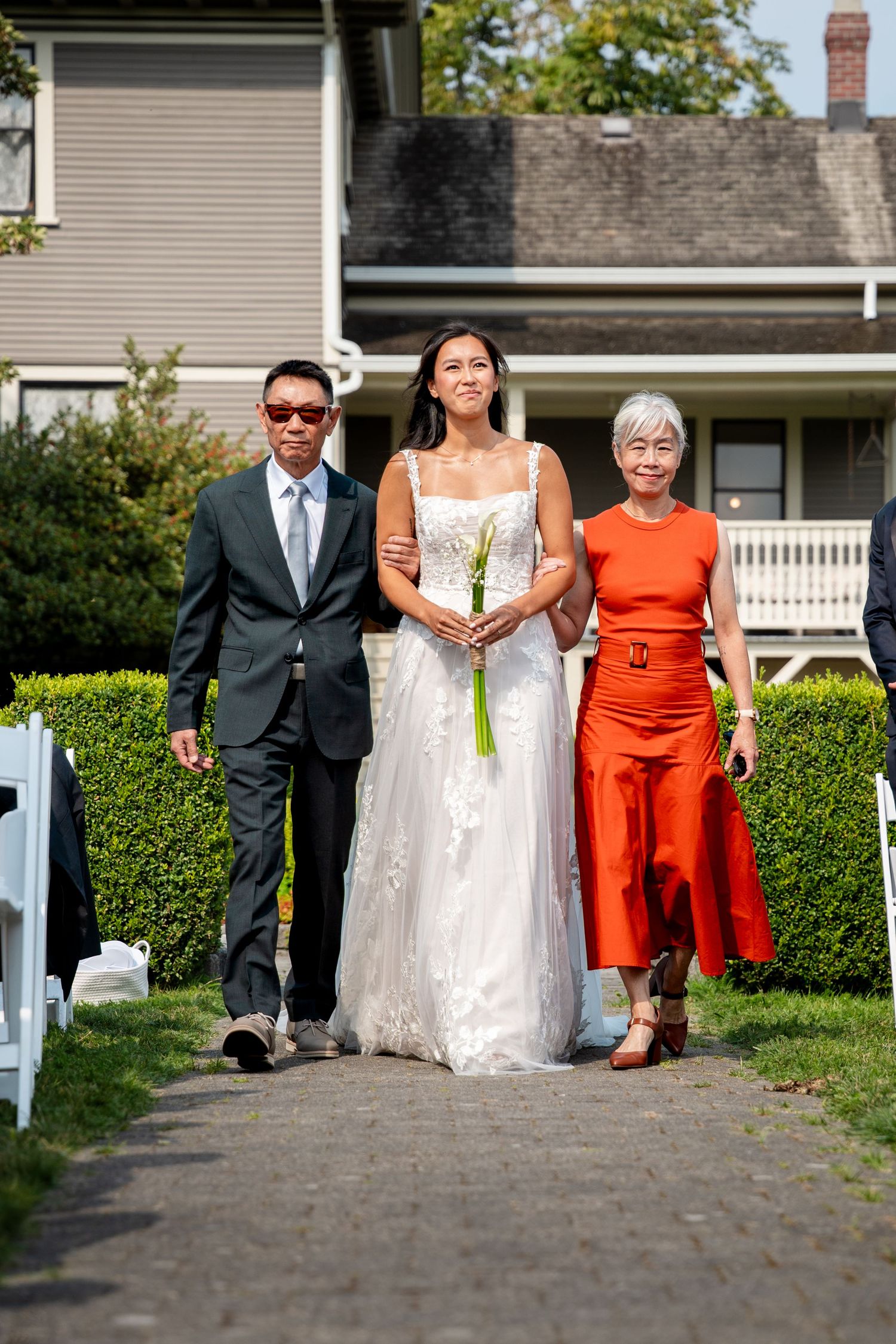 Bride walks down outdoor aisle with parents at sunny garden wedding ceremony near historic home.