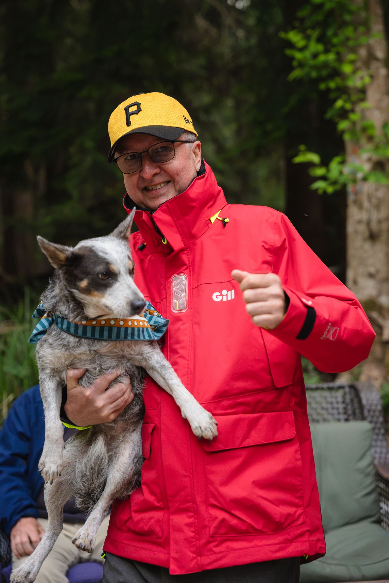 Smiling man in red Gill jacket and yellow Pittsburgh Pirates cap holding a blue heeler dog outdoors.